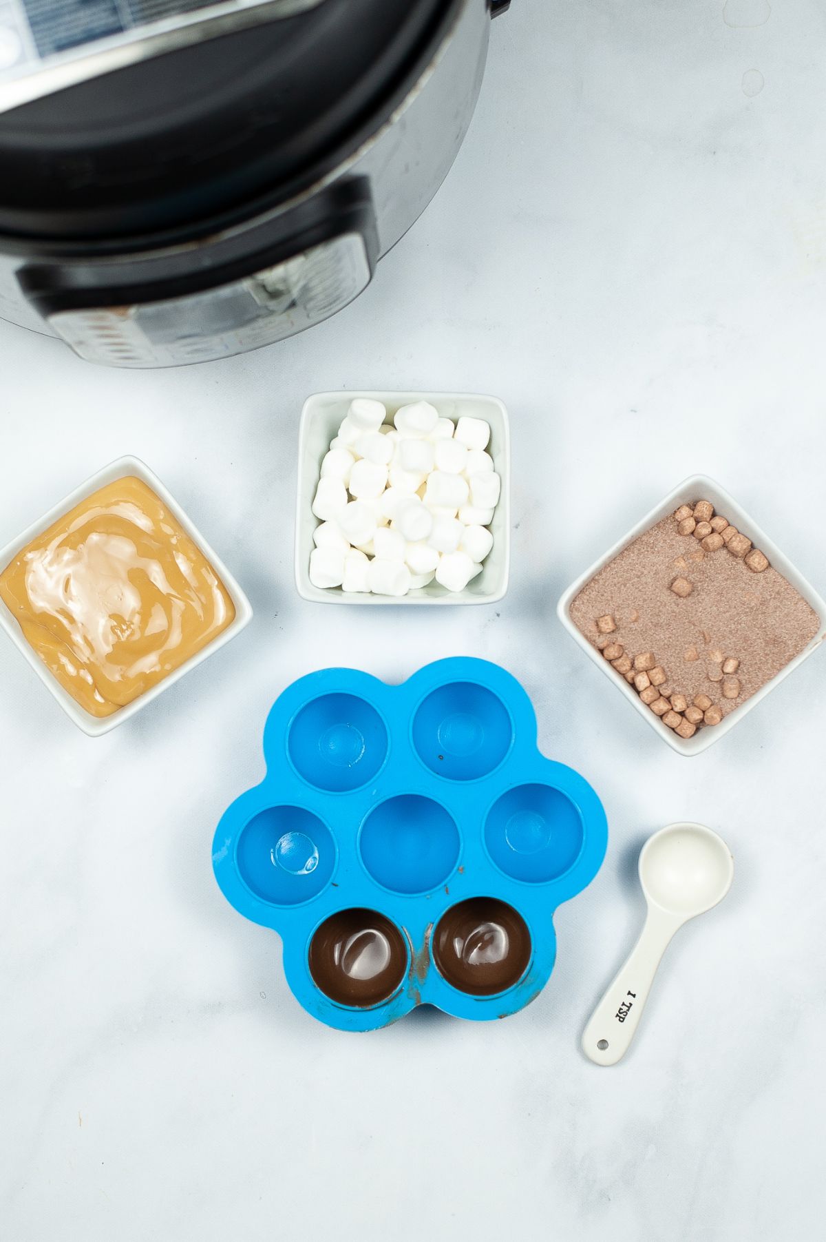 Chocolates in a silicone mold to form the chocolate bomb shell next to caramel sauce, marshmallows, and cocoa mix in white bowls.