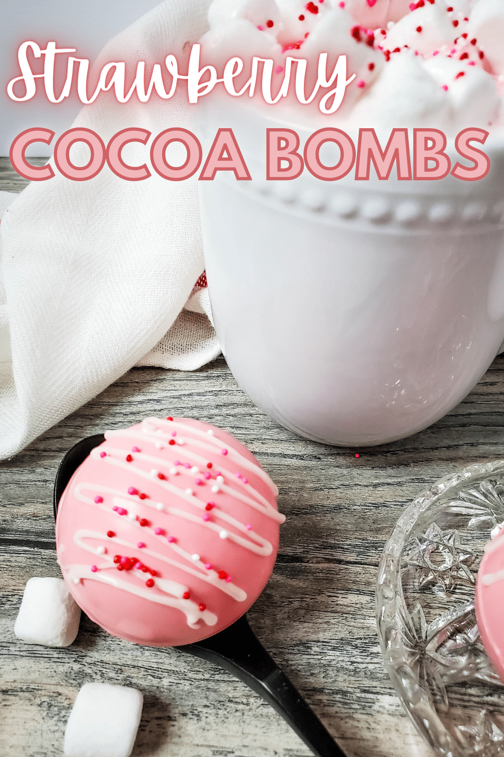 Strawberry Hot Cocoa Bombs on a spoon and a small glass plate next to a white mug of hot cocoa topped with marshmallows and red and pink sprinkles with a text at the upper part of the image saying Strawberry Cocoa Bombs.