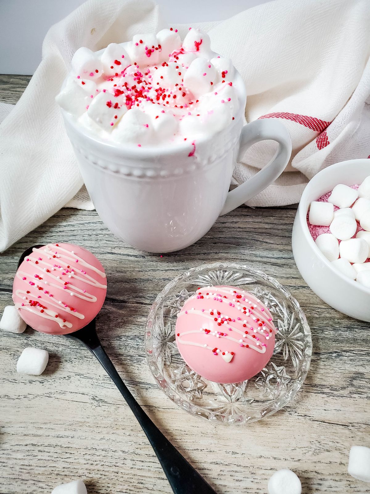 Strawberry Hot Cocoa Bombs on a small glass plate and on a spoon. Above them is a white cup  of hot cocoa with marshmallow toppings.