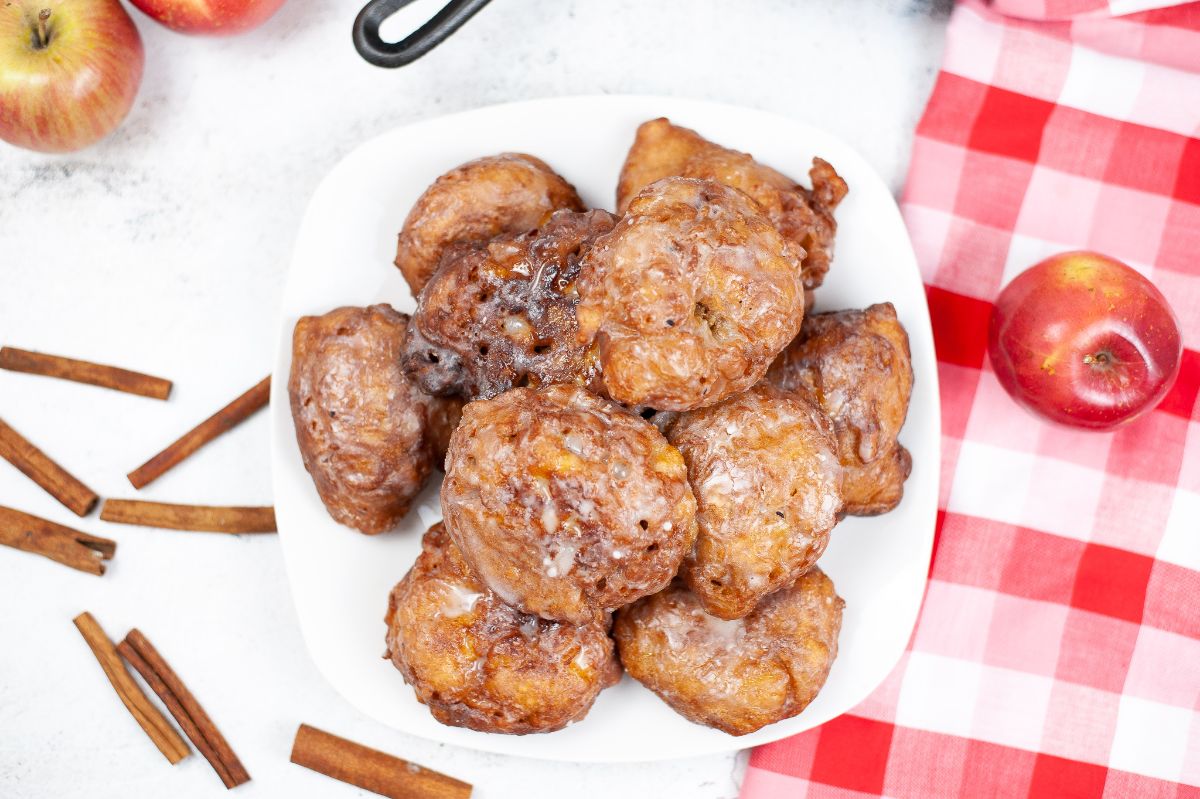 Overhead horizontal shot of Apple Fritters on a white serving plate. next to cinnamon sticks and an apple on a red and white checkered cloth