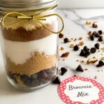 A beautifully layered brownie mix in a jar, adorned with a golden bow, features rich chocolate chips, brown sugar, white sugar, cocoa, and flour. A charming label reads "Brownie Mix," while scattered chocolate chips nearby invite indulgence.