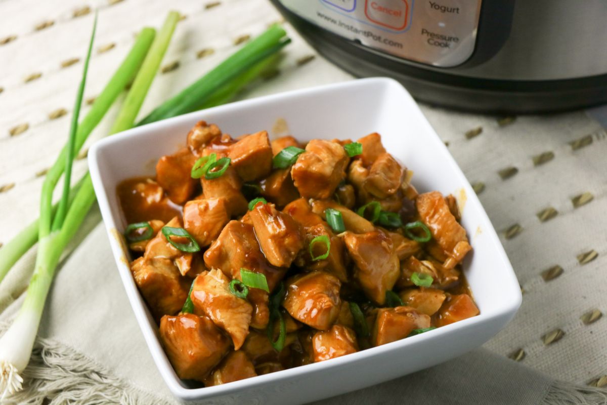 Horizontal image of Instant Pot General Tso's Chicken in a white serving bowl next to an instant pot.