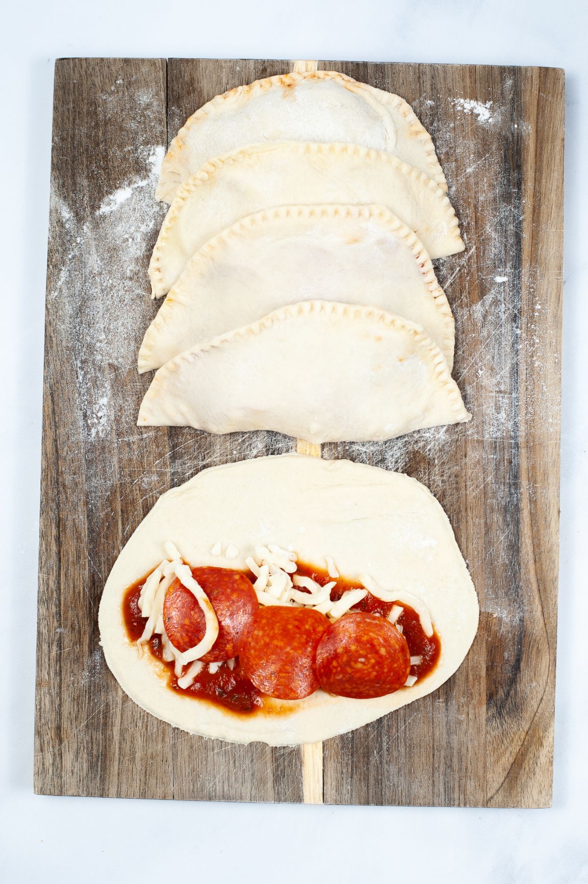4 assembled calzones on top of a wooden board with the 5th one still open, showing the pizza sauce, cheese, and pepperoni filling.