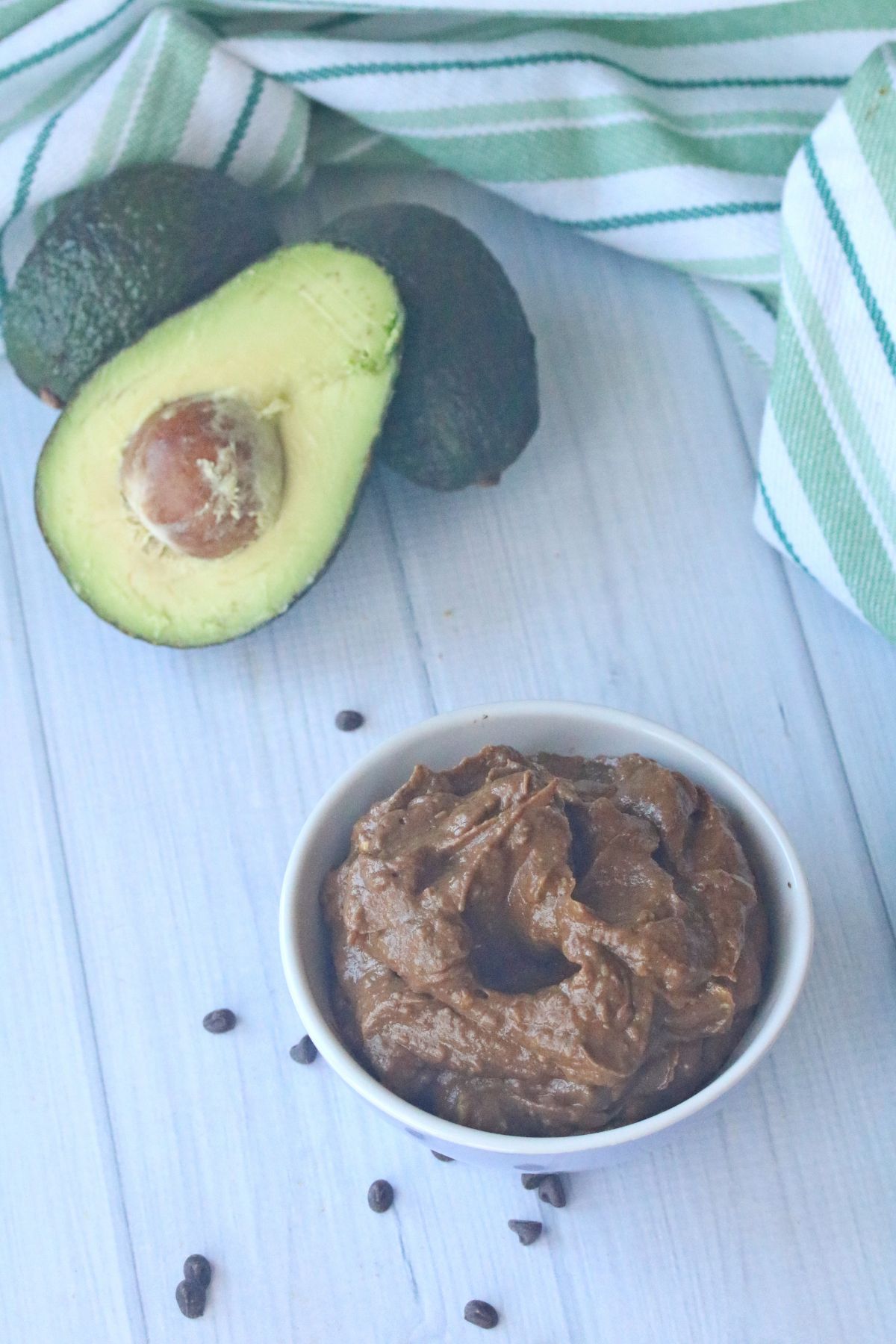 Vertical image of Chocolate Avocado Mousse in a white serving bowl at the lower right part of the image and fresh avocados at the upper left part of the image.