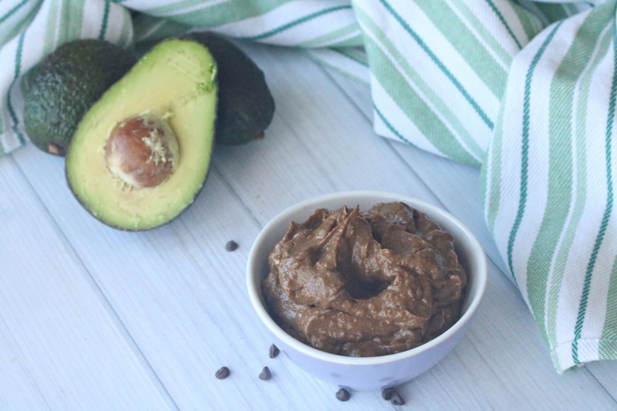 Horizontal image: Chocolate Avocado Mousse in a white serving bowl at the lower right part of the image and fresh avocados at the upper left part of the image.