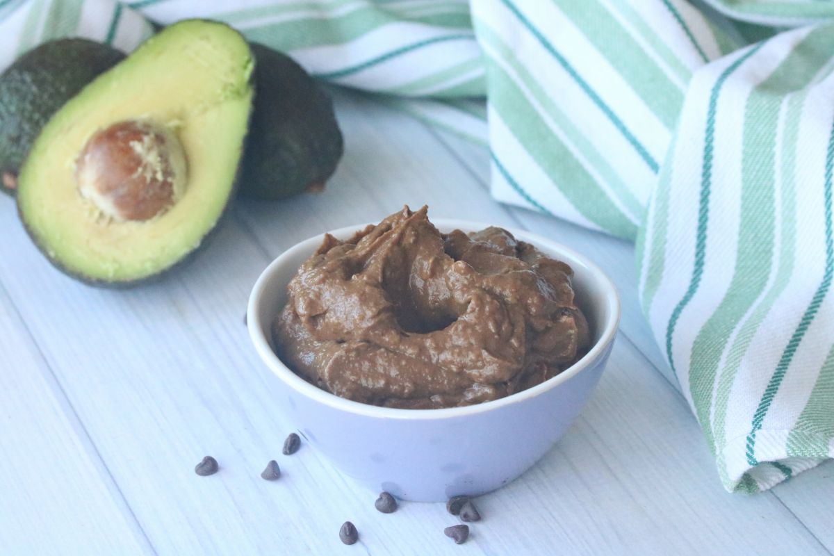 Horizontal image: Chocolate Avocado Mousse in a white serving bowl at the lower right part of the image and fresh avocados at the upper left part of the image.
