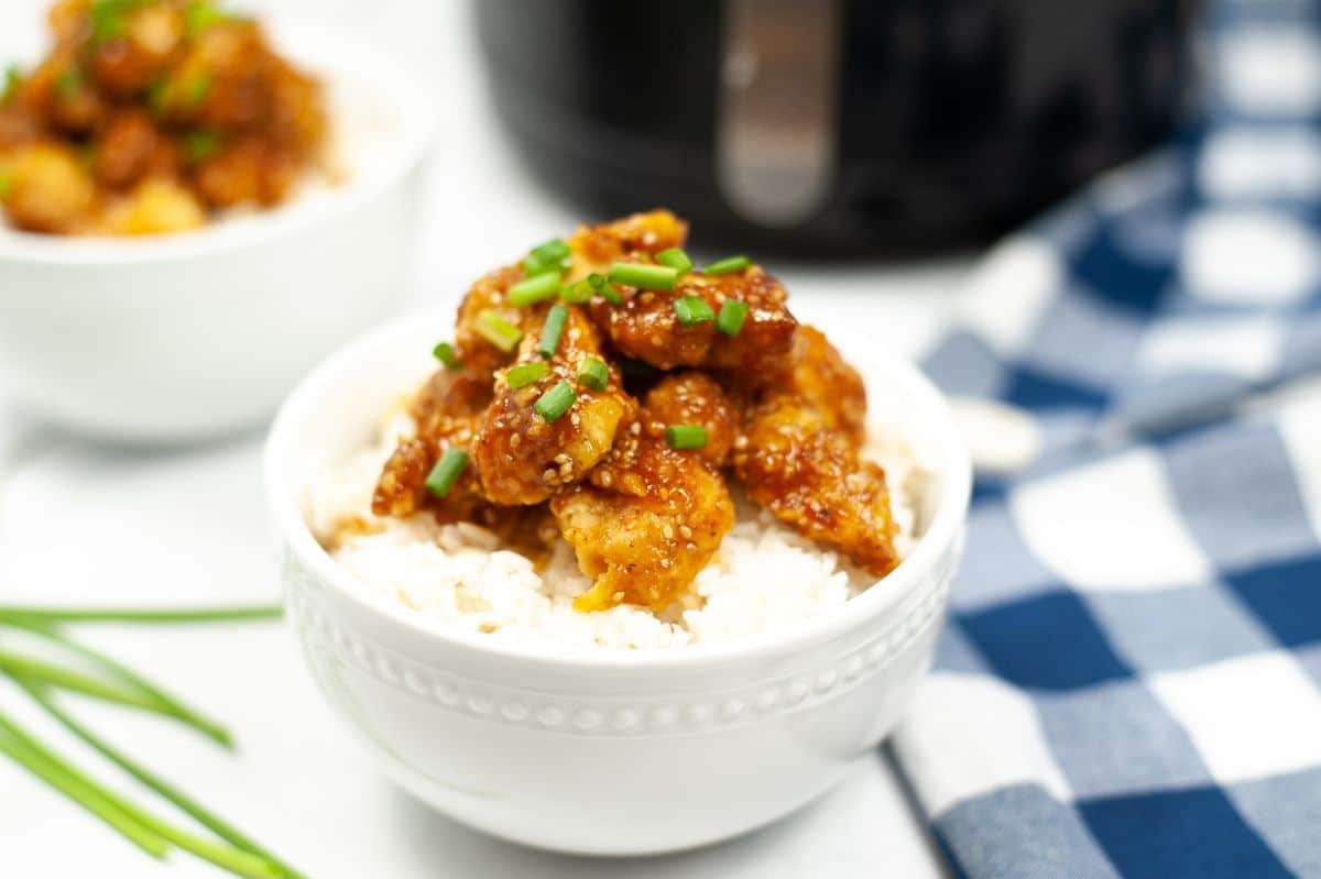 Air Fryer Sesame Chicken in a white bowl topped with spring onion with an air fryer in the background