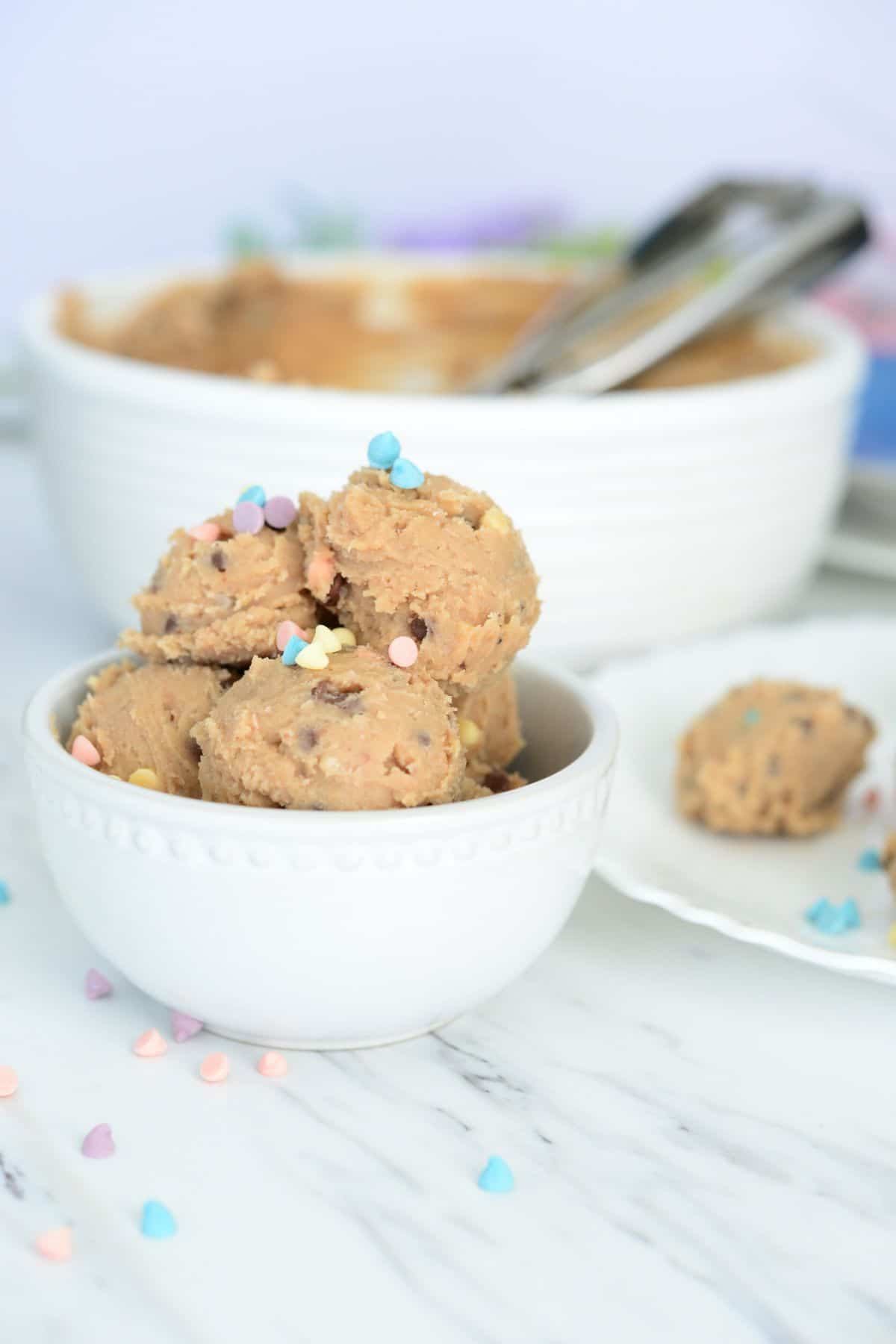 No Bake Cookie Dough Bites in a white bowl topped with pastel color sprinkles with a bowl of dough blurred in the background