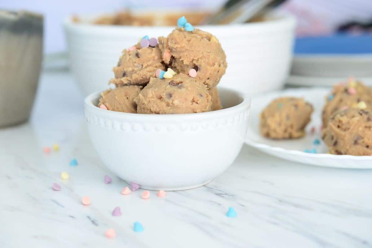No Bake Cookie Dough Bites in a white bowl topped with pastel color sprinkles with a bowl of dough blurred in the background