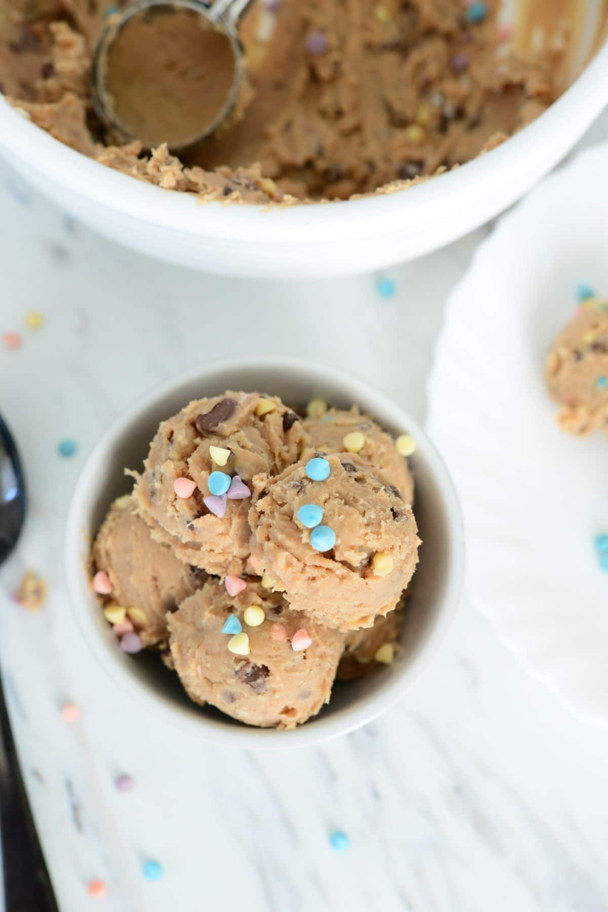 overhead shot of No Bake Cookie Dough Bites in a white bowl topped with pastel color sprinkles with a bowl of dough blurred in the background