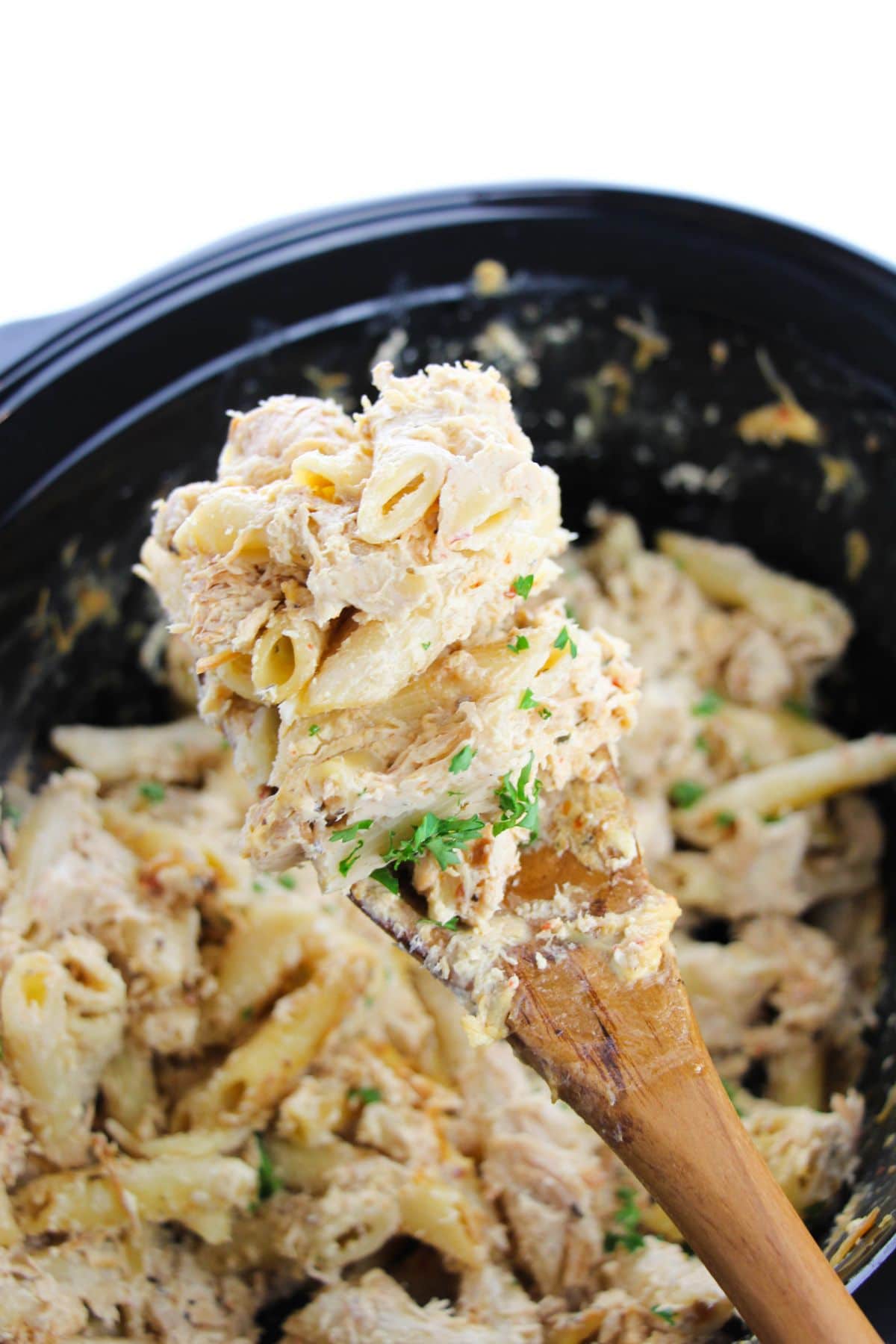 A portion of Olive Garden Crock Pot Chicken on a wooden spoon with more food in a crock pot under it.