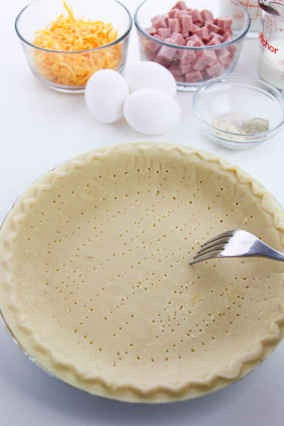 a fork being used to prick holes in a Pie crust with the other quiche ingredients in the background