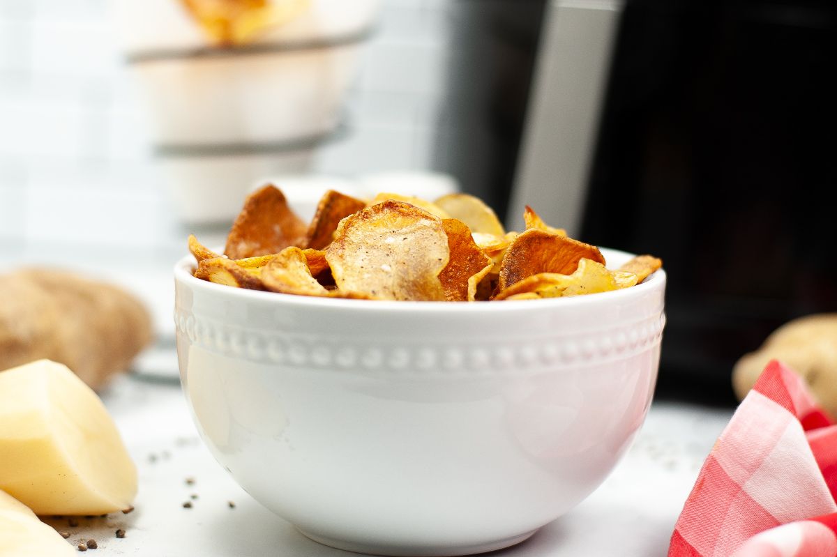 Air fryer potato chips in a white bowl next to potatoes and a red and white cloth.