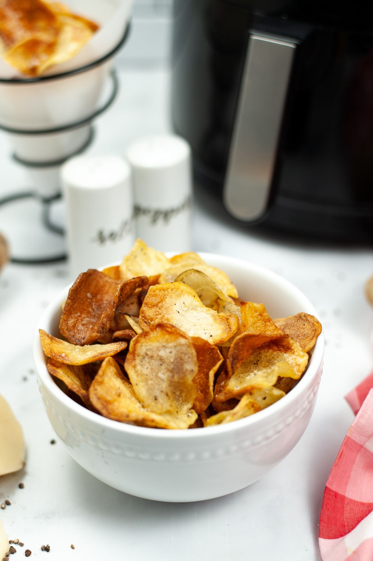 Homemade Air Fryer Potato Chips in white bowl with salt and pepper and an air fryer blurred in the background