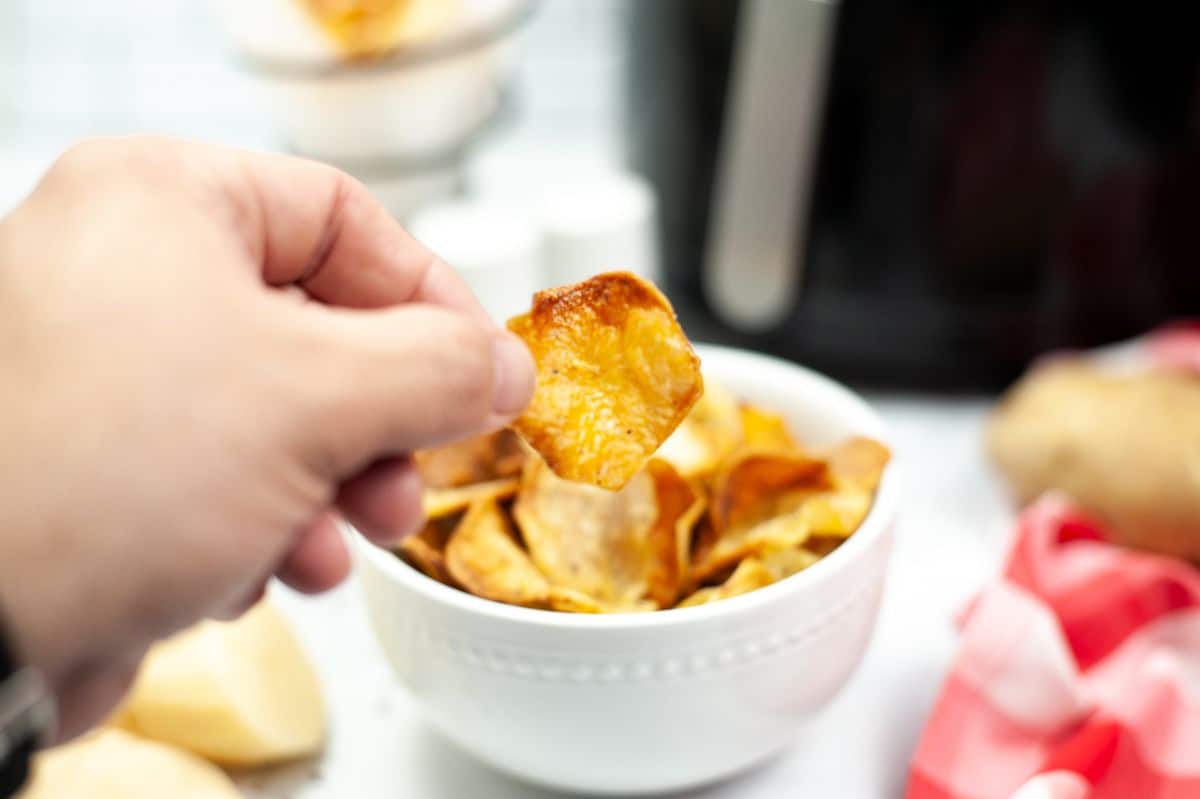 a hand holding a potato chip with the rest of the Homemade Air Fryer Potato Chips in white bowl 