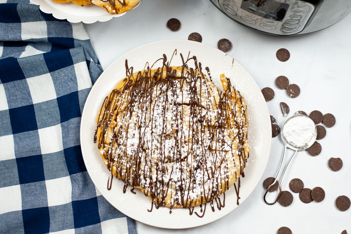 overhead view of Homemade Funnel Cake on a white plate dusted with powder sugar and drizzled with chocolate with an instant pot and more funnel cakes on a white cake stand in the background