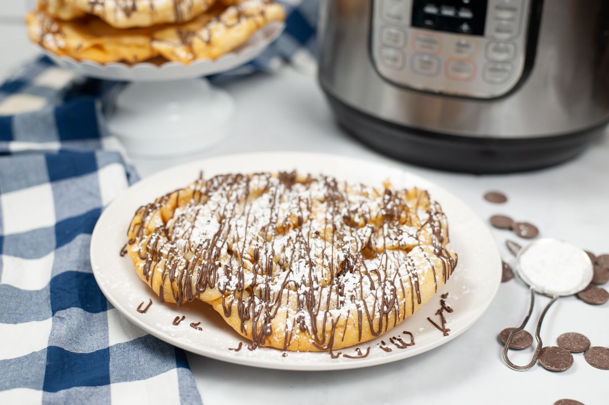 Homemade Funnel Cake on a white plate dusted with powder sugar and drizzled with chocolate with an instant pot and more funnel cakes on a white cake stand in the background