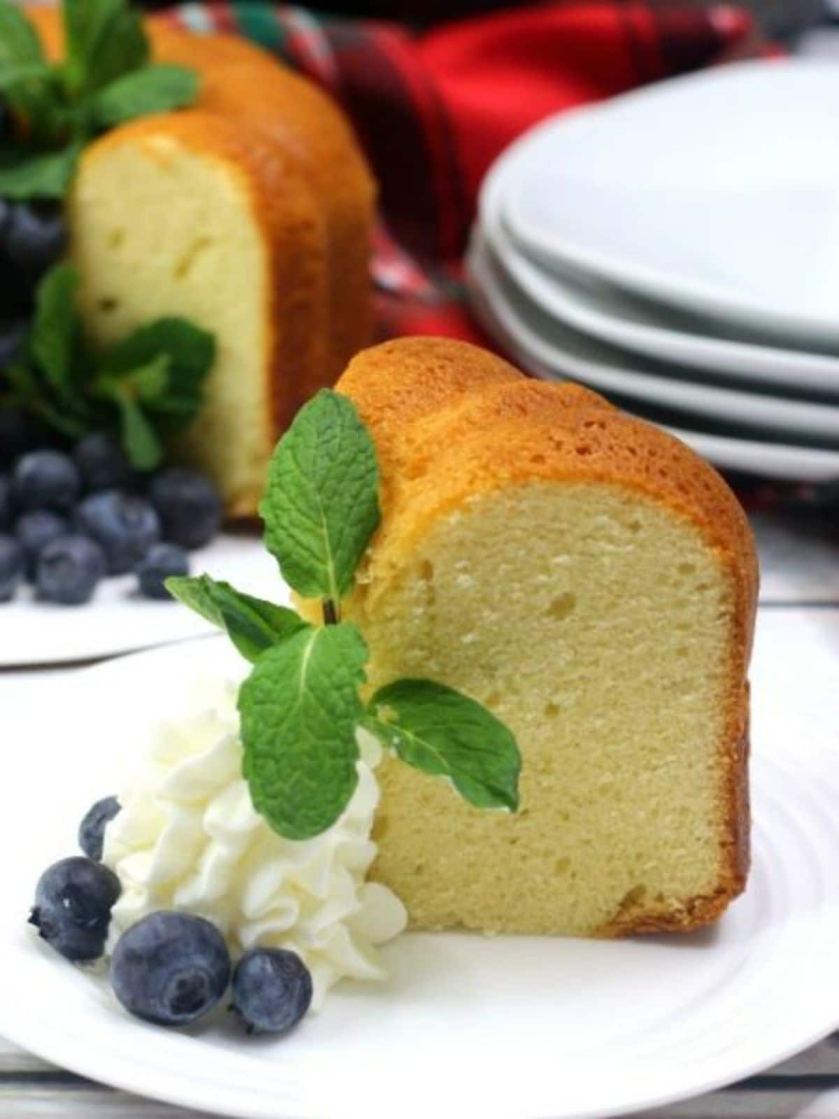 a slice of instant pot pound cake with whipped cream, a mint leaf, and blueberries on a white plate with the rest of the cake, more blueberries and a stack of white plates blurred in the background