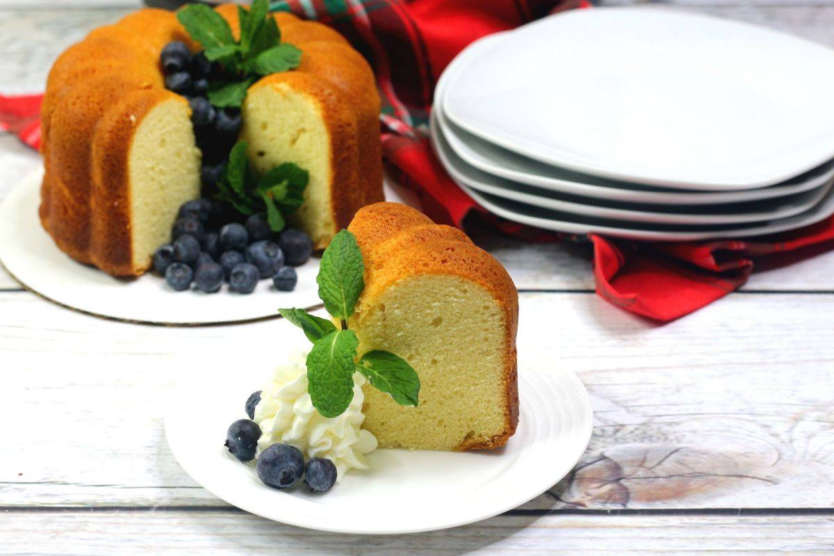 a slice of instant pot pound cake with whipped cream, a mint leaf, and blueberries on a white plate with the rest of the cake, more blueberries and a stack of white plates in the background
