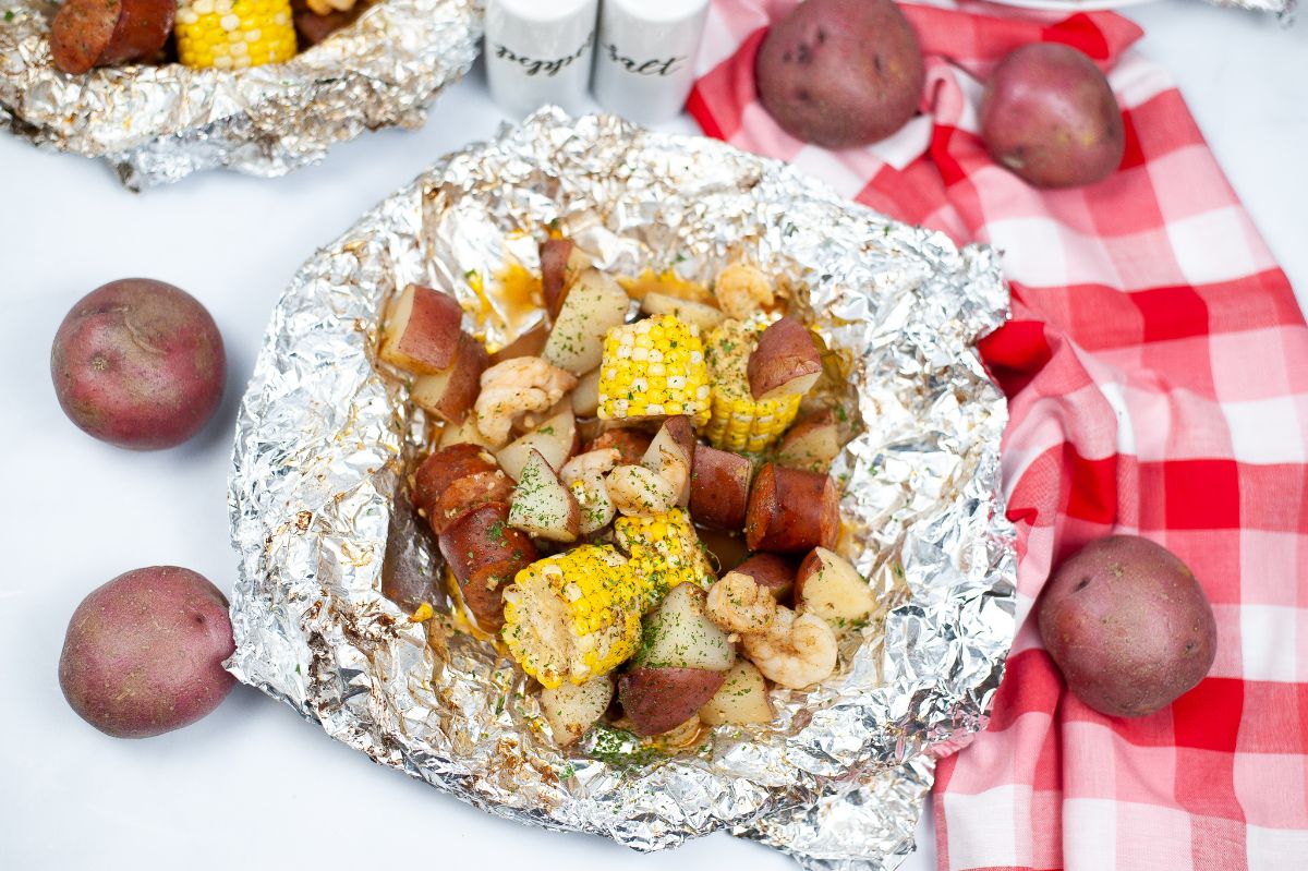 Low Country Boil Foil Packets next to red potatoes, a red and white checkered cloth, and salt and pepper shakers