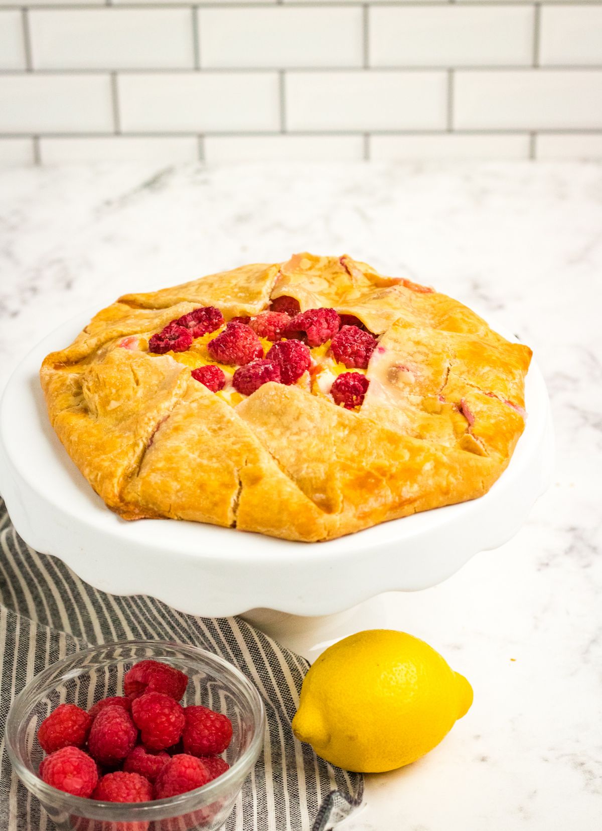 Raspberry Cream Cheese Galette on a white cake stand next to a striped cloth, a lemon and raspberries in a glass bowl