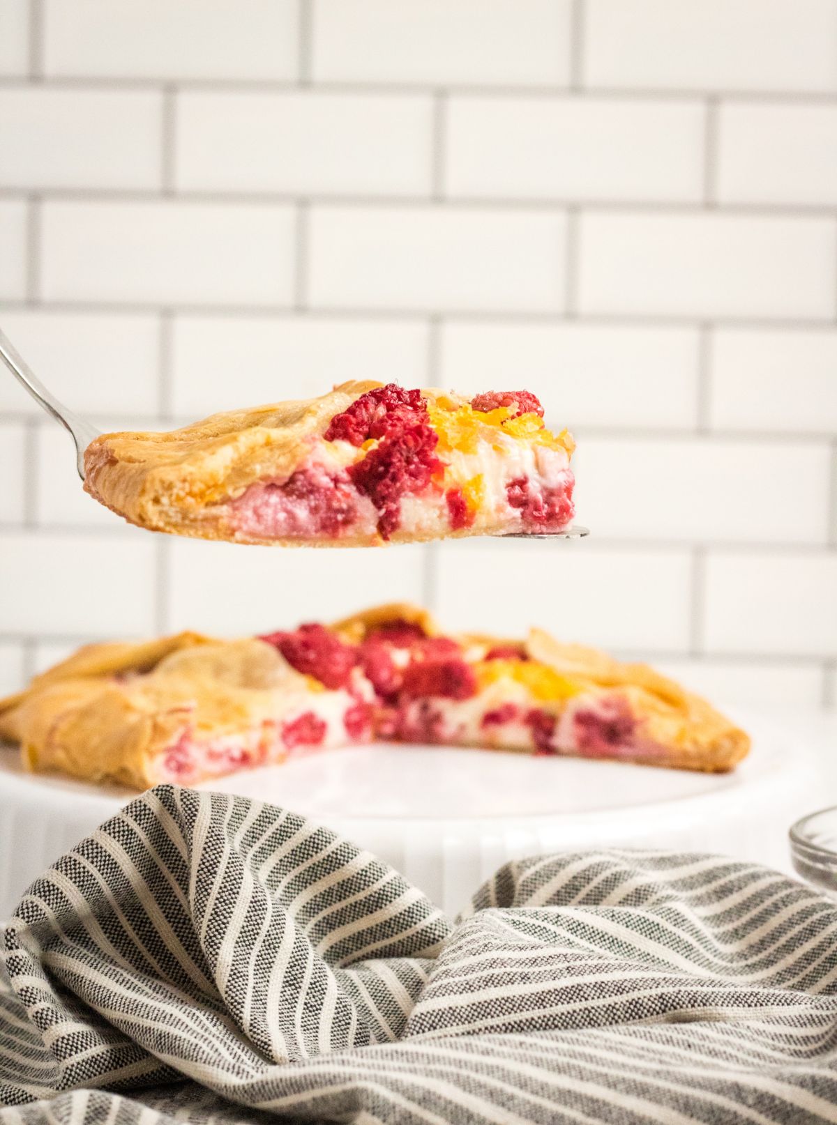 a closeup of a slice of Raspberry Cream Cheese Galette on a spatula above a striped cloth, with more of the dessert in the background