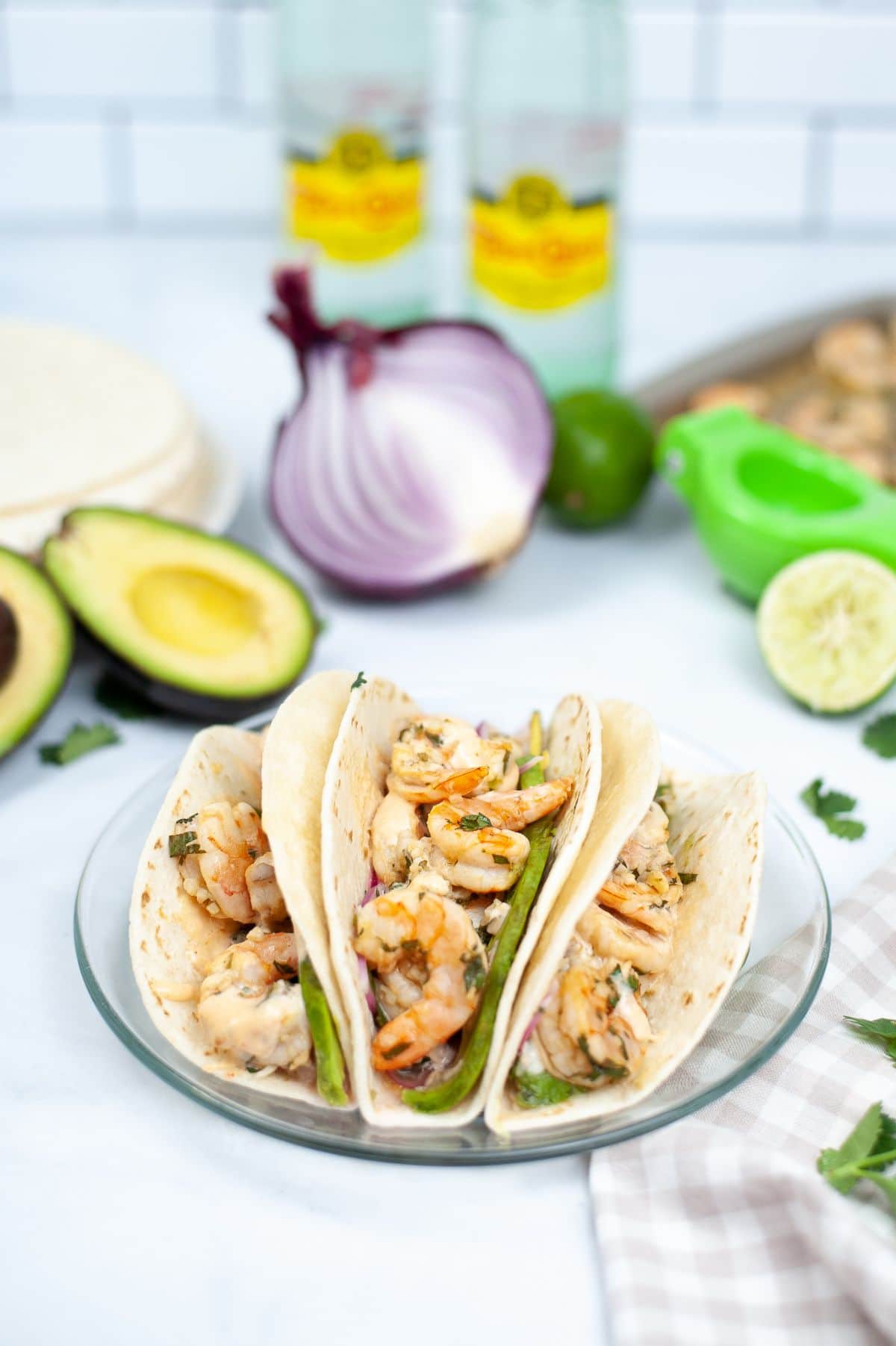 Sheet Pan Shrimp Tacos on a glass plate with an avocado, onion and limes blurred in the background