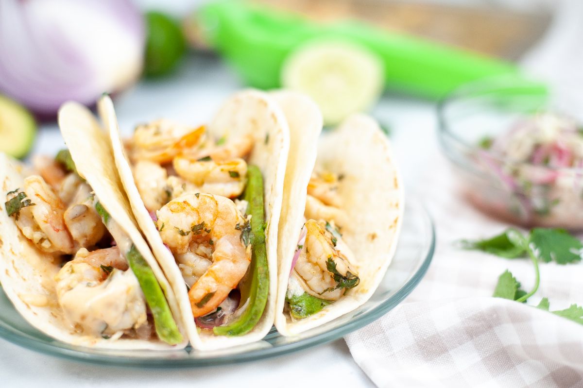 Sheet Pan Shrimp Tacos on a glass plate with an avocado, onion and limes blurred in the background