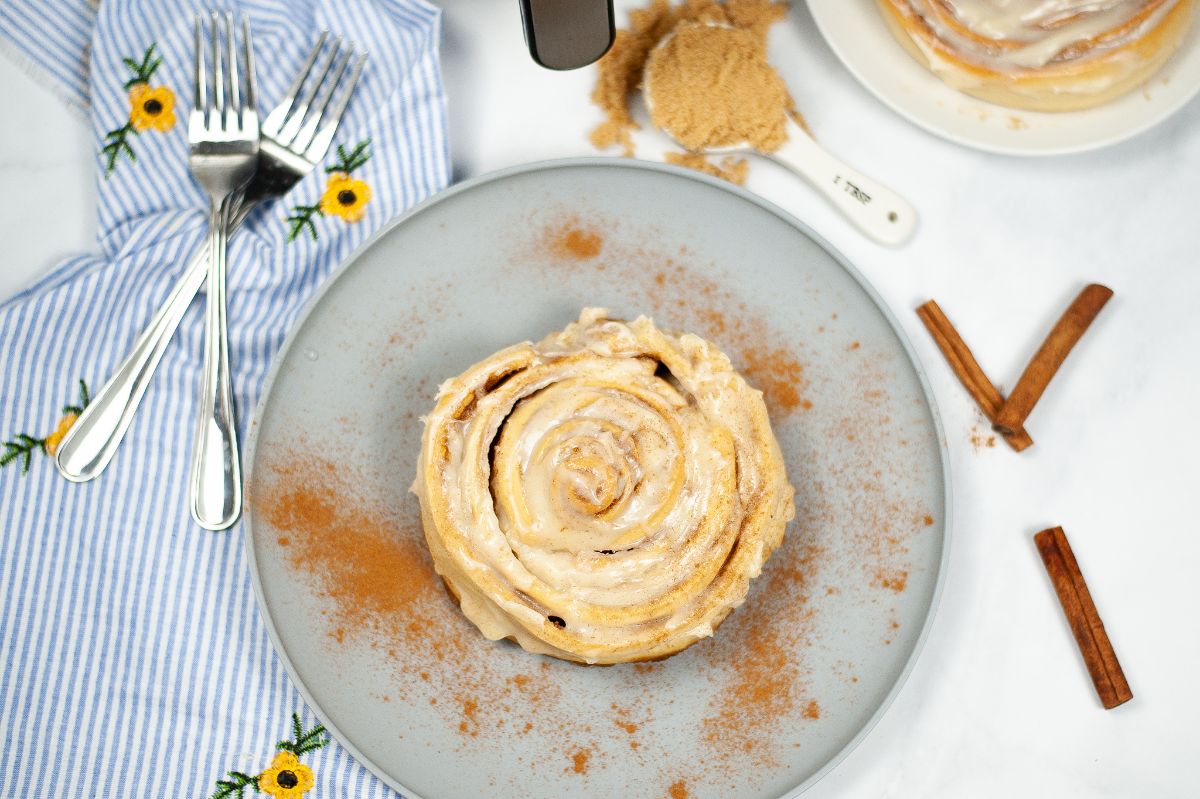 overhead view of an Air Fryer Copycat Cinnabon Cinnamon Roll topped with cream and dusted with cinnabon powder next to forks a blue and white striped cloth, brown sugar in a tablespoon and cinnamon sticks