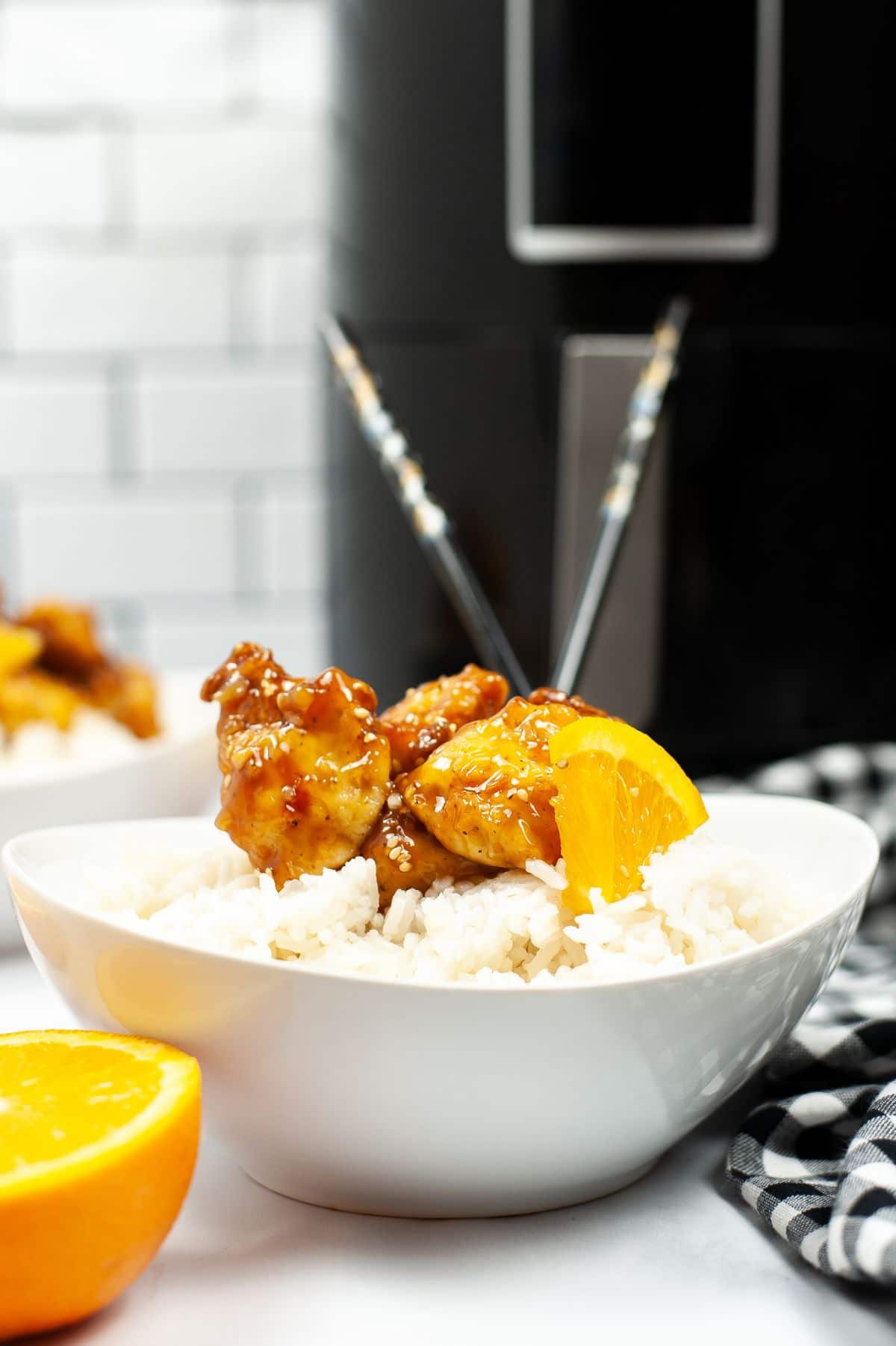 Air Fryer Copycat Panda Express Orange Chicken in a white bowl with rice  next to a halved orange with an air fryer an another bowl of chicken blurred in the background
