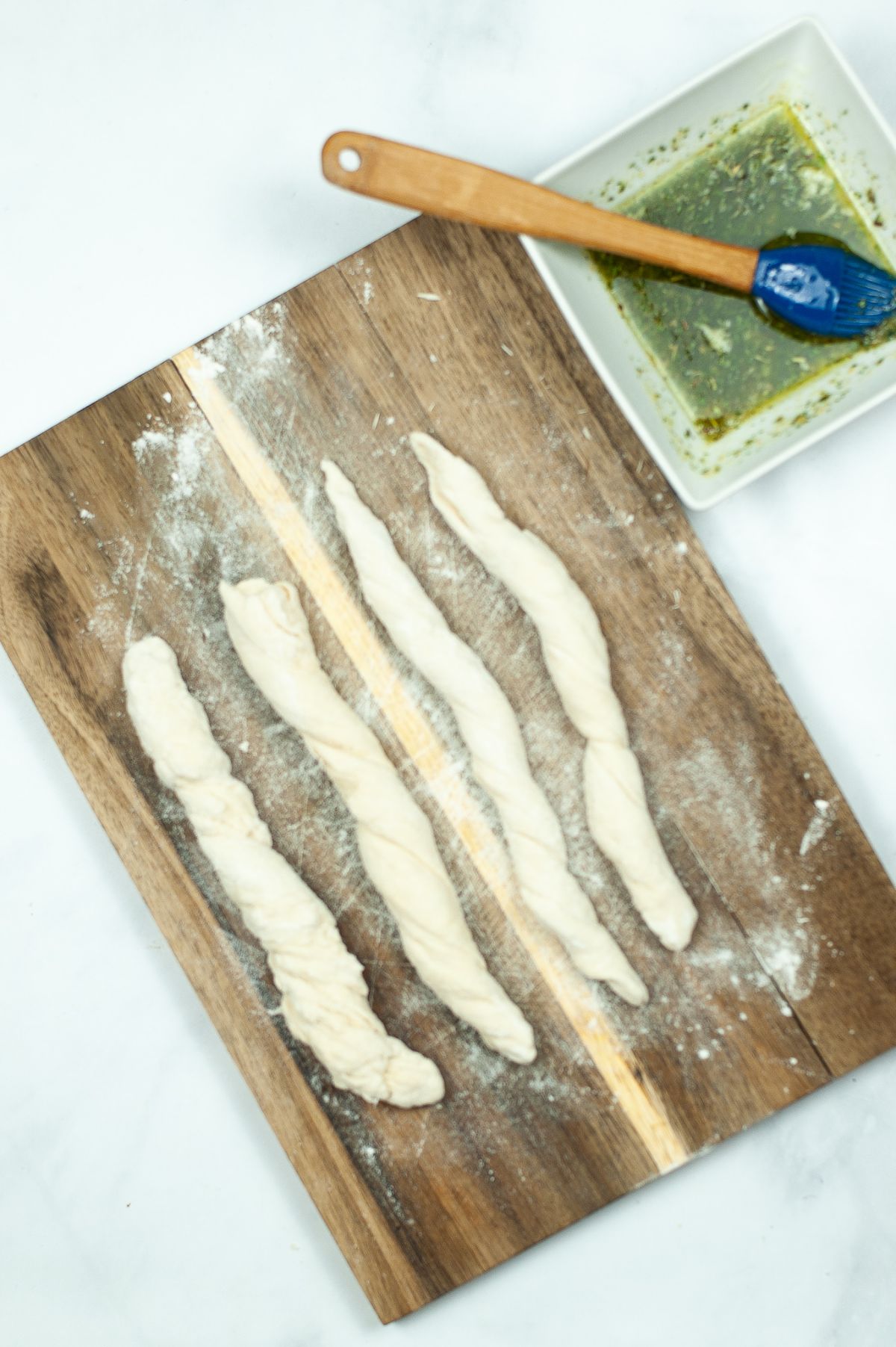 Twisted dough to make breadsticks on a wooden board next to melted butter and herbs in a white bowl with a pastry brush