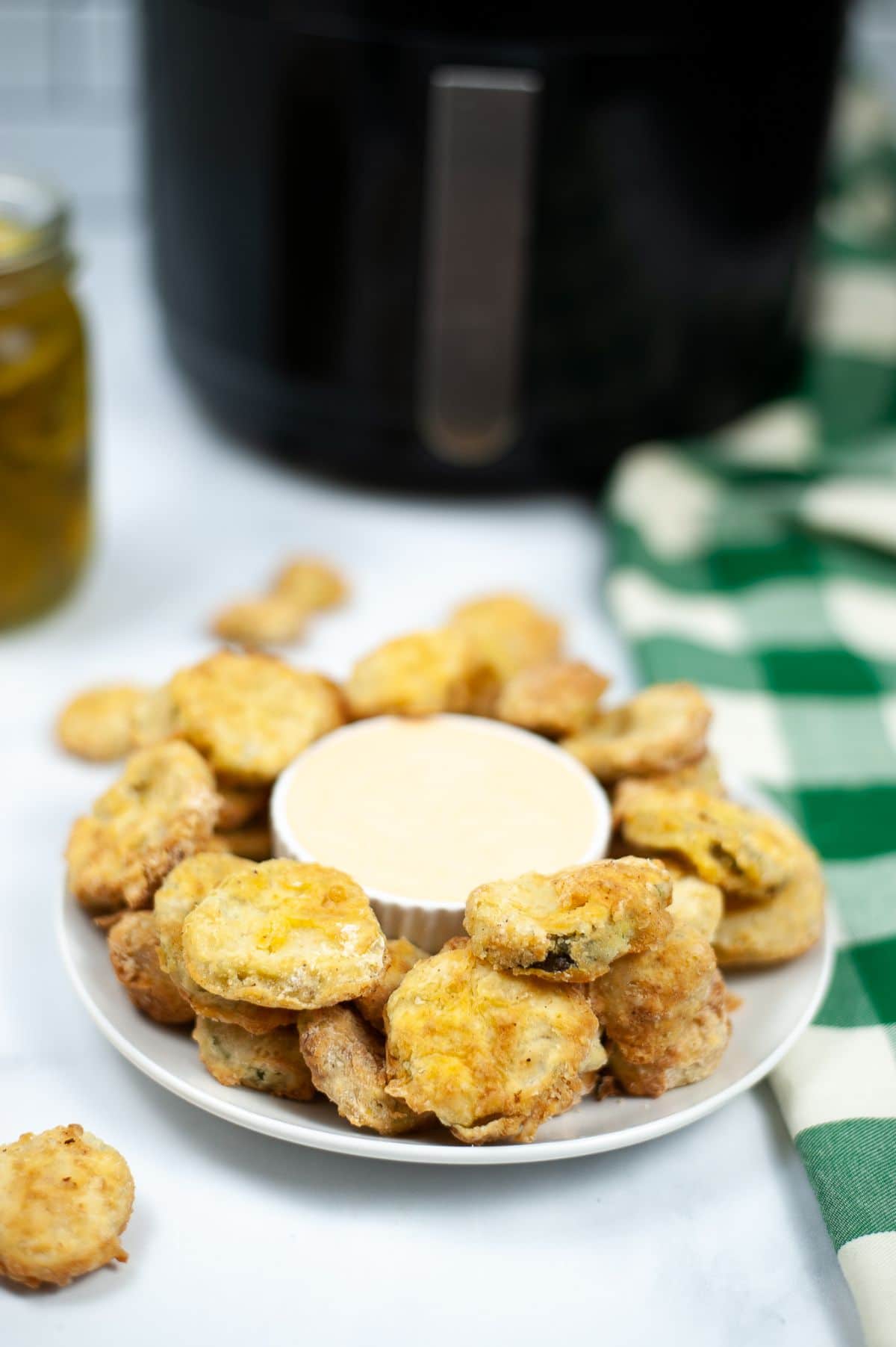 Air Fryer Pickles on a white plate with dipping sauce in the middle with a jar of pickles, an air fryer and a green and white checkered cloth blurred in the background