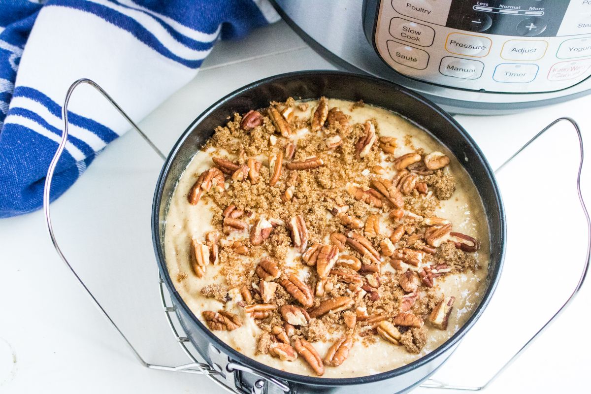 Applesauce Cake mixture in a pan topped with pecan nuts next to an instant pot and a blue and white cloth