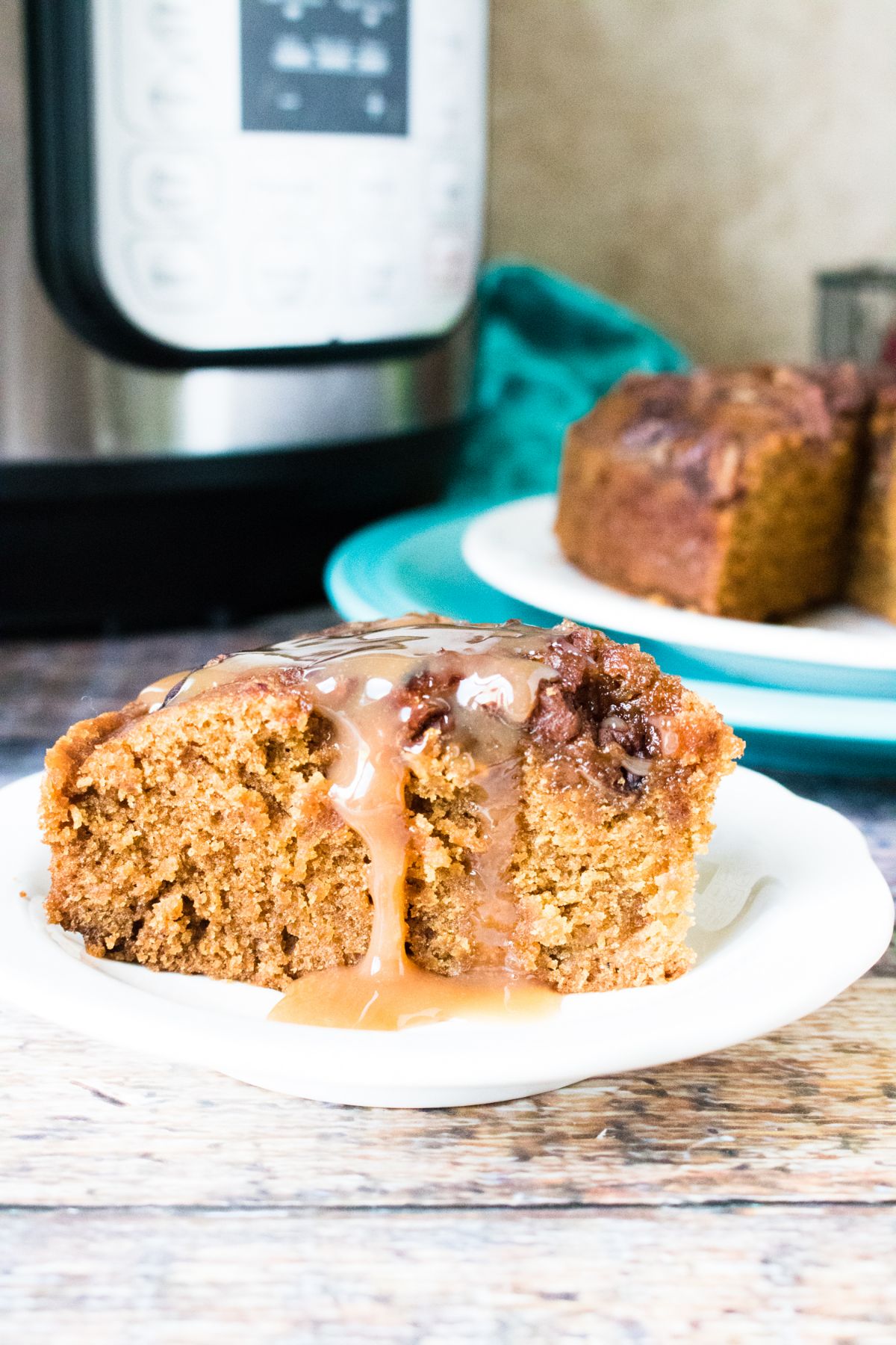 a slice of Instant Pot Applesauce Cake on a white plate topped with caramel sauce with an instant pot and more cake on a plate in the background
