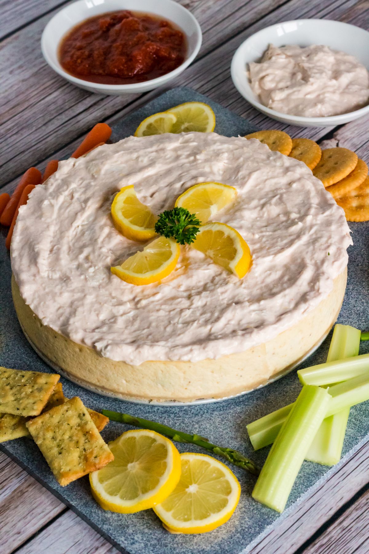 Instant Pot Crab Cheesecake on a square stone plate topped with lemon and parsley, with  celery, carrots, asparagus, lemon and crackers on the side.