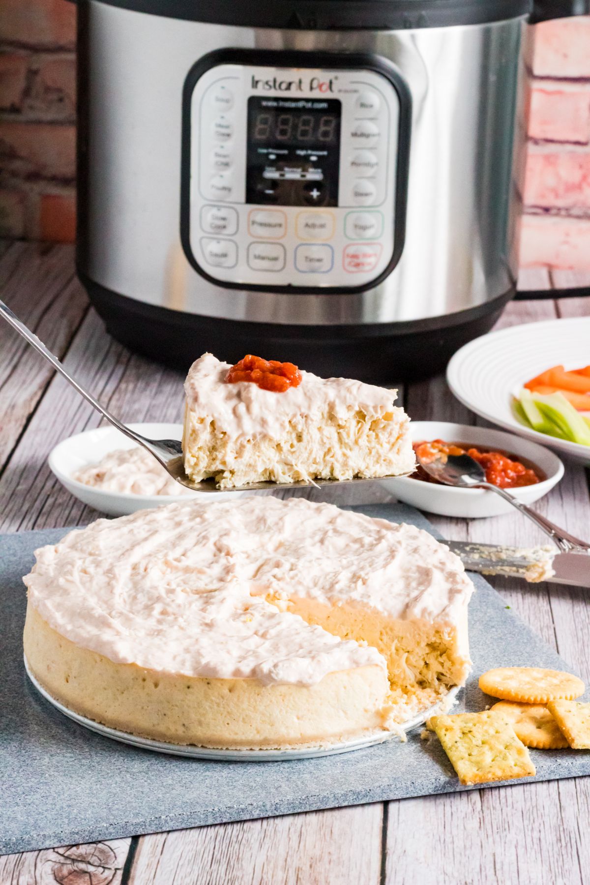 Instant Pot Crab Cheesecake on a round metal plate on a square stone cutting board with crackers on the side, with a piece of cheesecake on a spatula above it, with bowl of sauce, carrots and vegetables on a white plate and an instant pot in the background