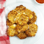A clear glass plate holds a stack of golden-brown, air-fried patties, revealing perfectly melted mozzarella slices inside. A red-checkered cloth is partially visible to the left and a small bowl with red sauce can be seen in the top right corner.