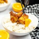 A bowl of white rice topped with copycat Panda Express orange chicken pieces, garnished with an orange slice. Chopsticks rest on the bowl, and a whole orange and checked cloth are in the background.