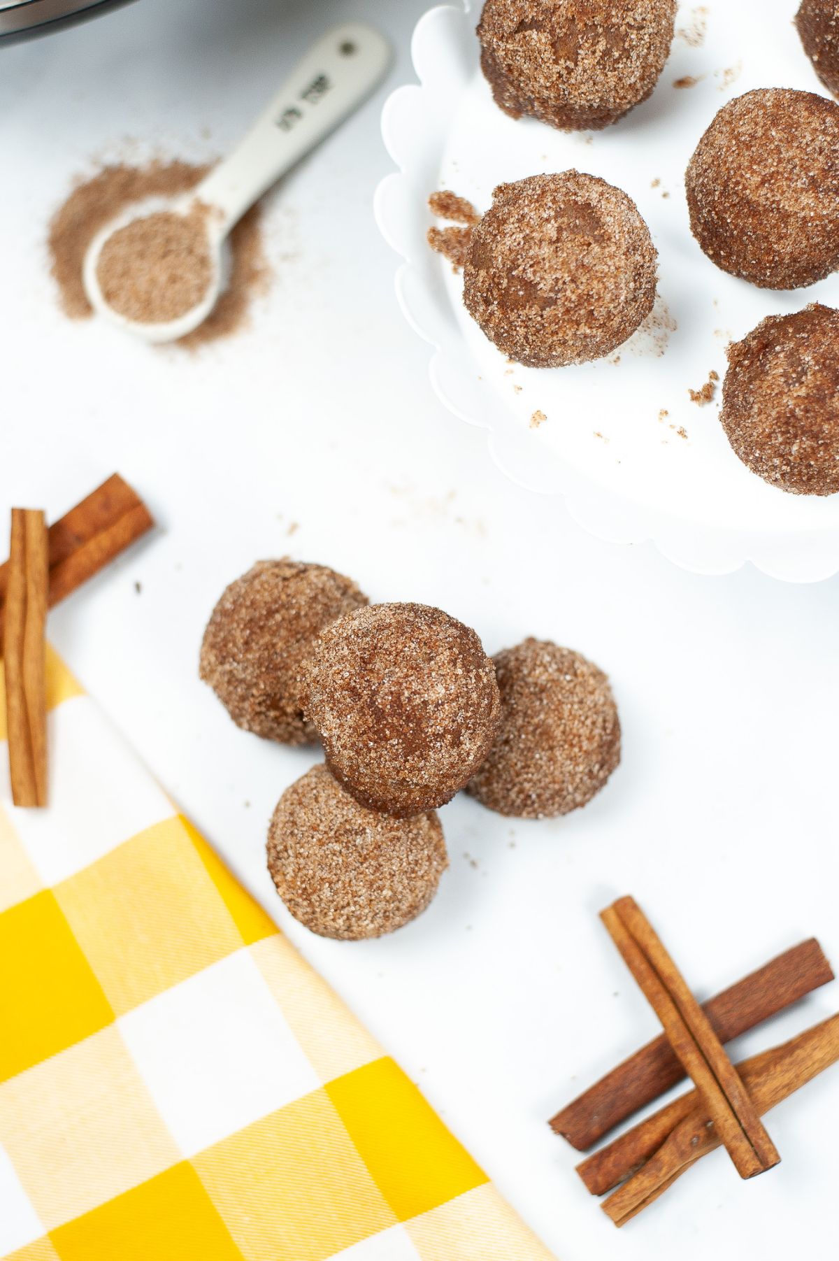 Instant Pot Churro Bites coated in cinnamon and sugar next to cinnamon sticks on a white plate on a yellow and white checkered cloth