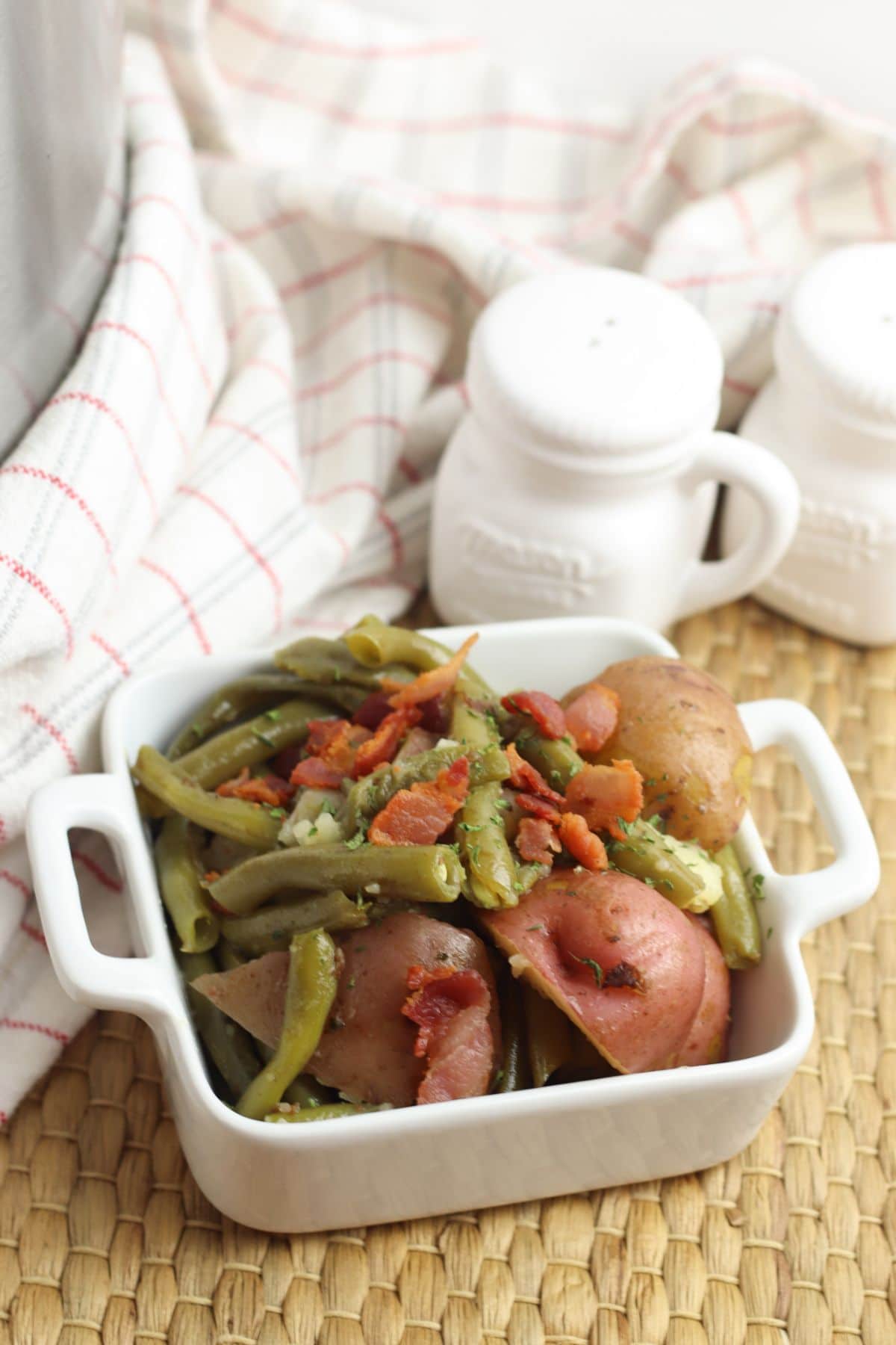 Instant Pot Green Beans and Potatoes topped with bacon in a square white dish next to a white cloth and white jars