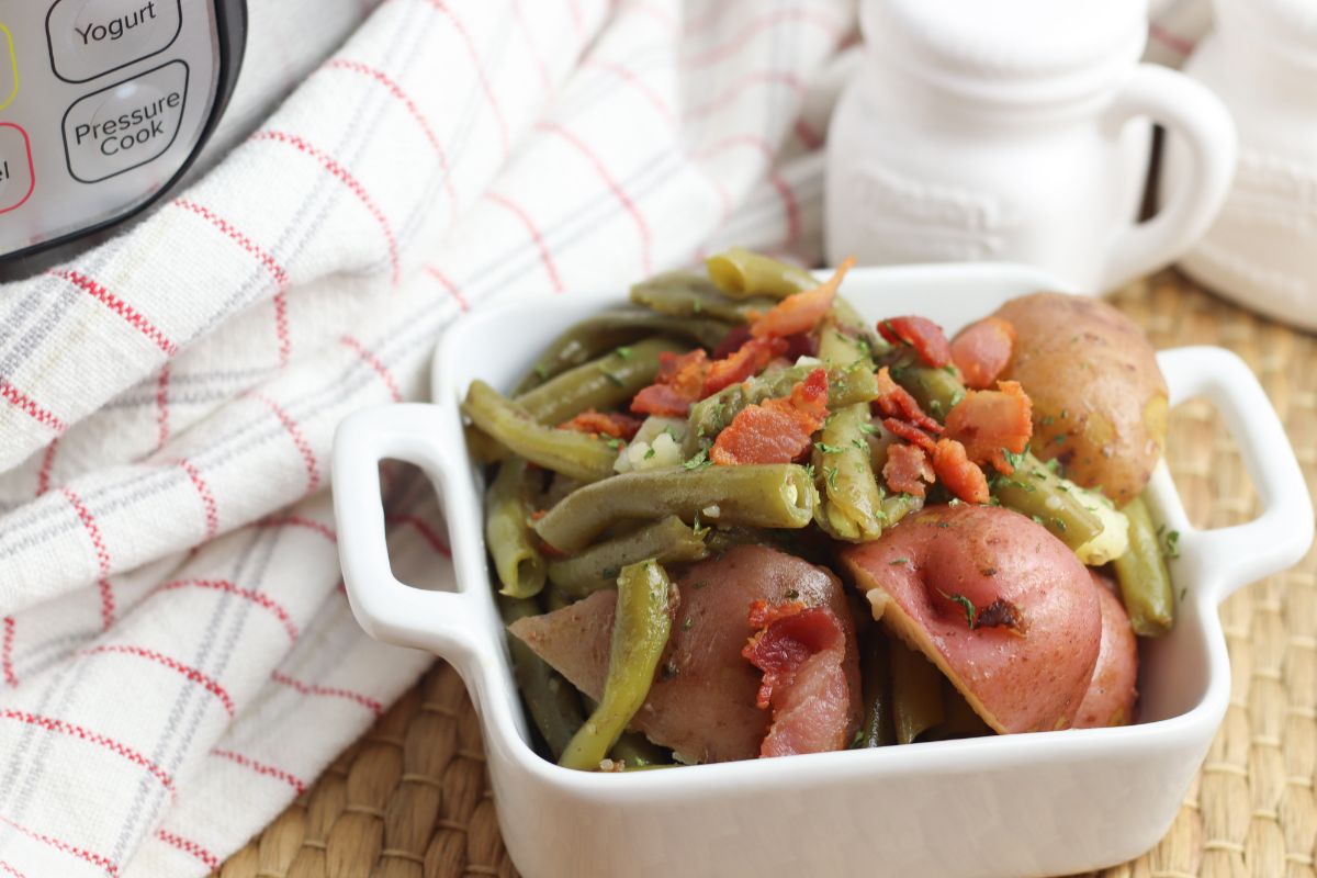 Instant Pot Green Beans and Potatoes topped with bacon in a square white dish next to a white cloth and white jars