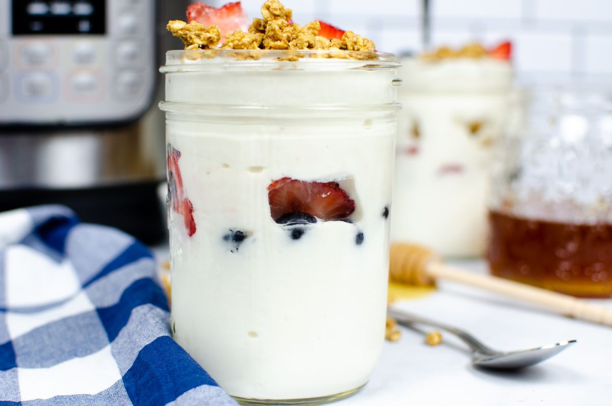Instant Pot Vanilla Yogurt in a jar topped with strawberry, blueberry and granola next to a blue and white checkered cloth with an instant pot blurred in the background