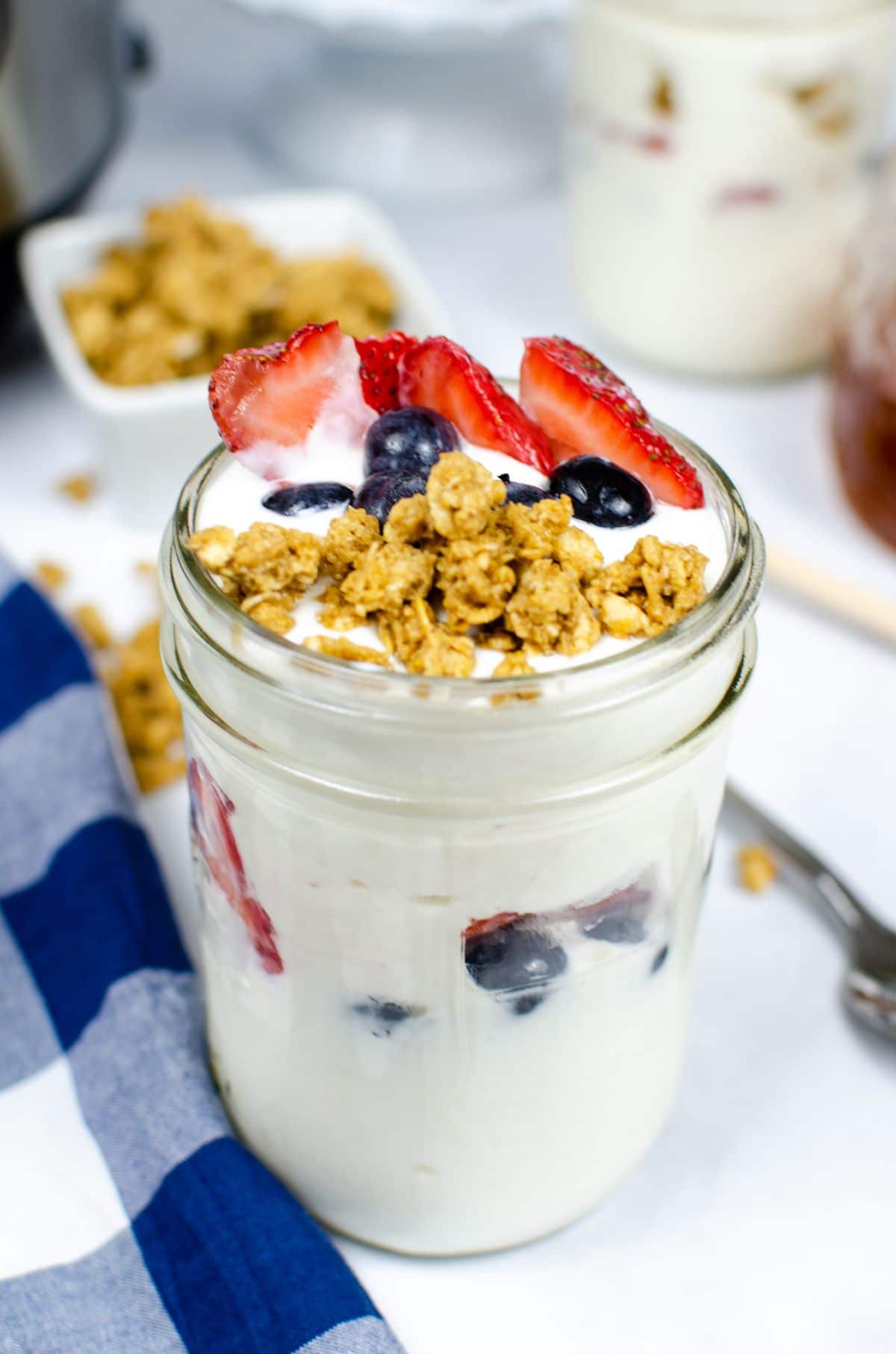 Instant Pot Vanilla Yogurt in a jar topped with strawberry, blueberry and granola next to a blue and white checkered cloth with an instant pot blurred in the background