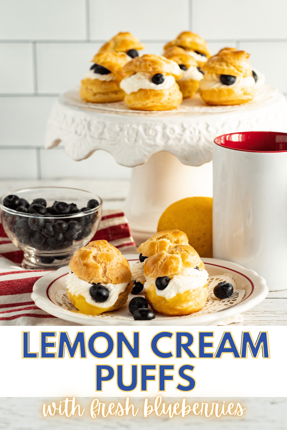 Lemon Cream Puffs With Fresh Blueberries on a plate and cake stand next to blueberries in a glass bowl, a lemon, a white and red mug and a white and red striped cloth with title text reading Lemon Cream Puffs with fresh blueberries