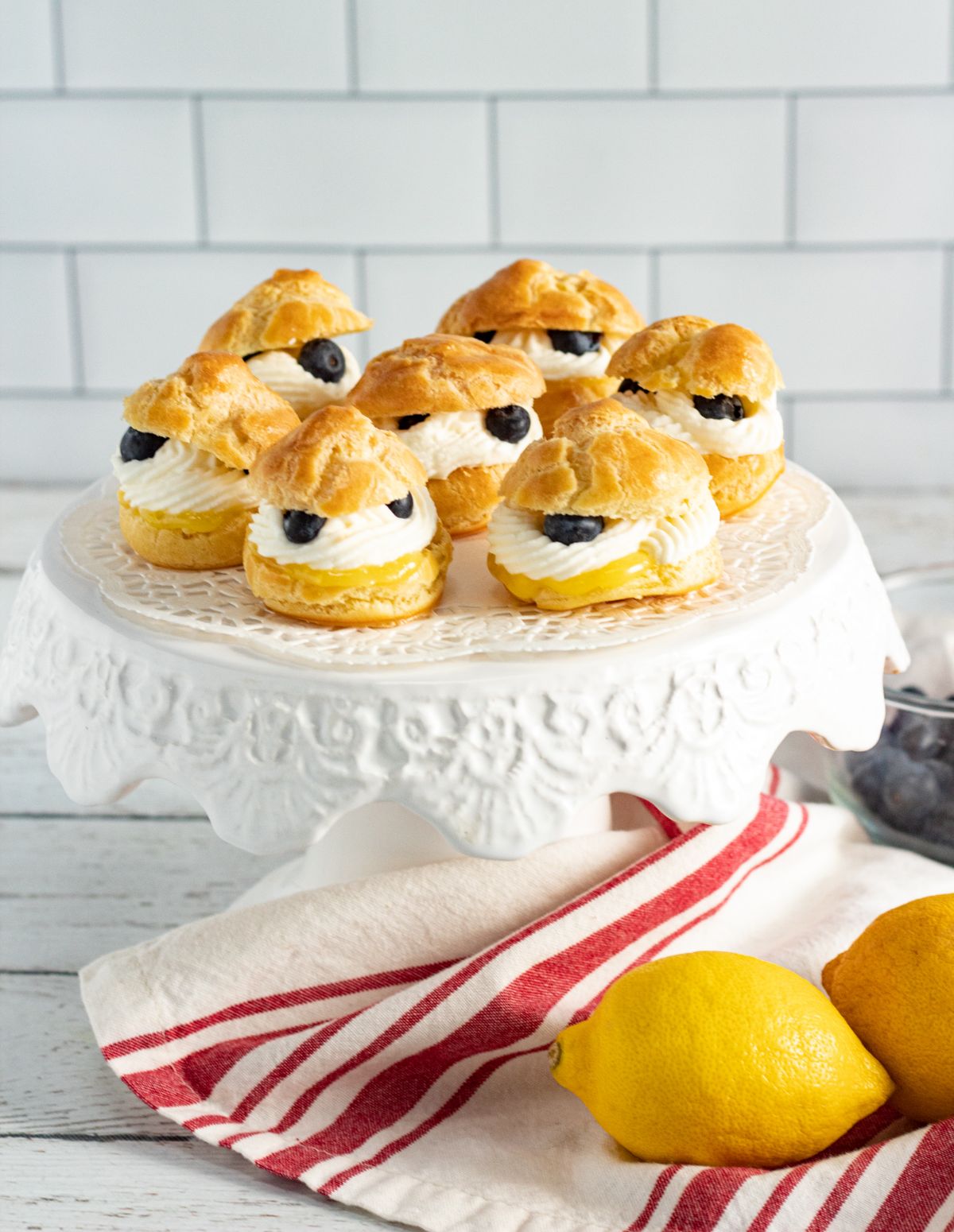 Lemon Cream Puffs With Fresh Blueberries on the cake stand next to a white and red striped cloth and two lemons