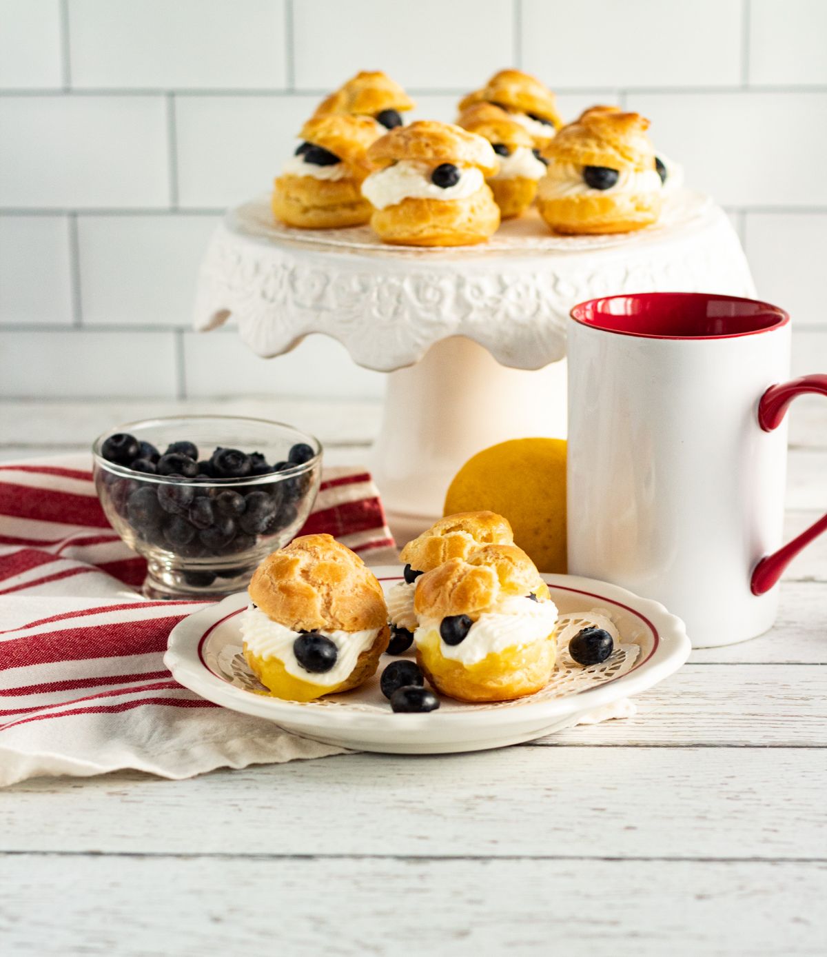 Lemon Cream Puffs With Fresh Blueberries on a plate and cake stand next to blueberries in a glass bowl, a lemon, a white and red mug and a white and red striped cloth