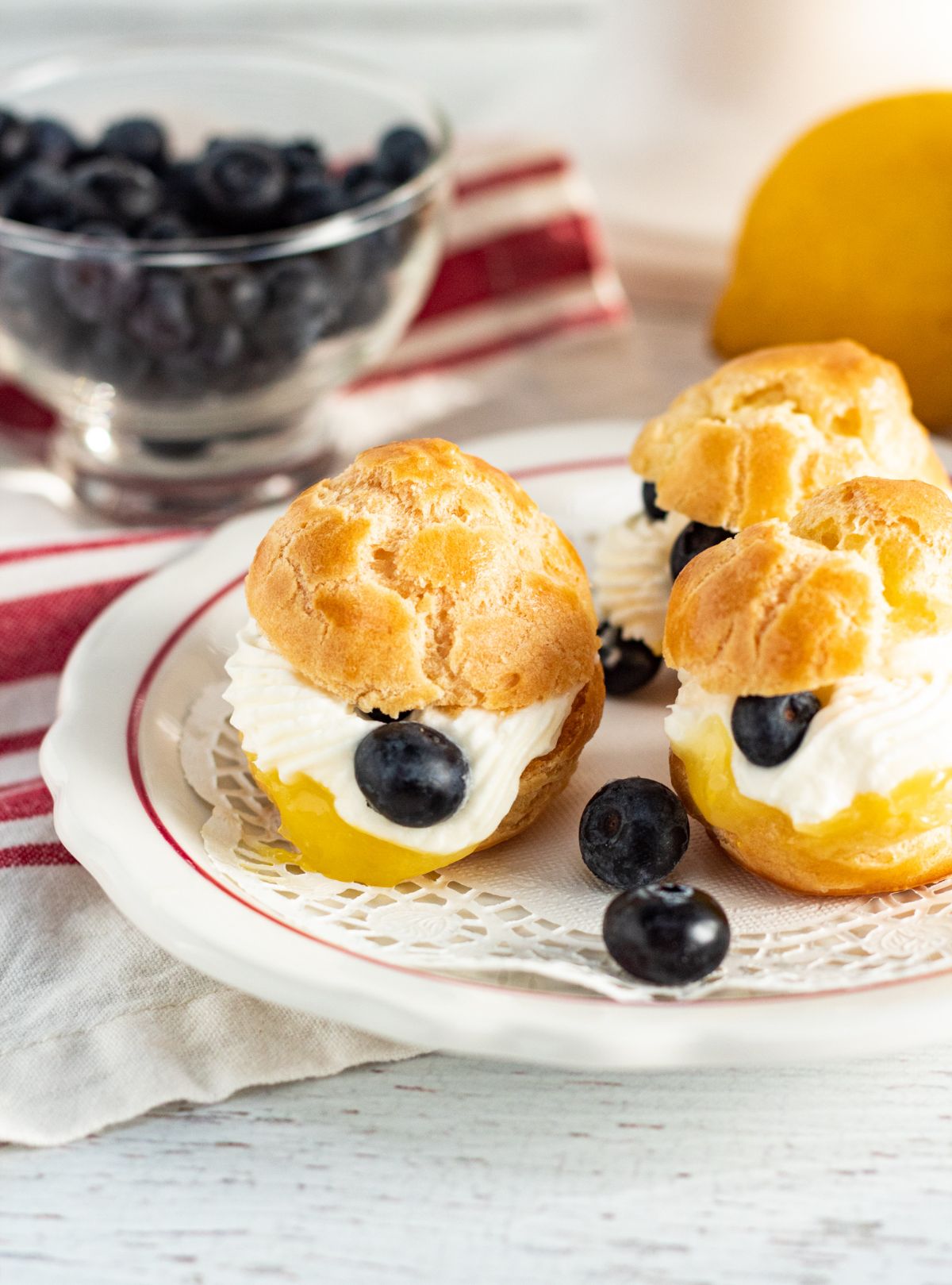 a closeup of Lemon Cream Puffs With Fresh Blueberries on a plate with blueberries in a glass bowl and a lemon blurred in the background
