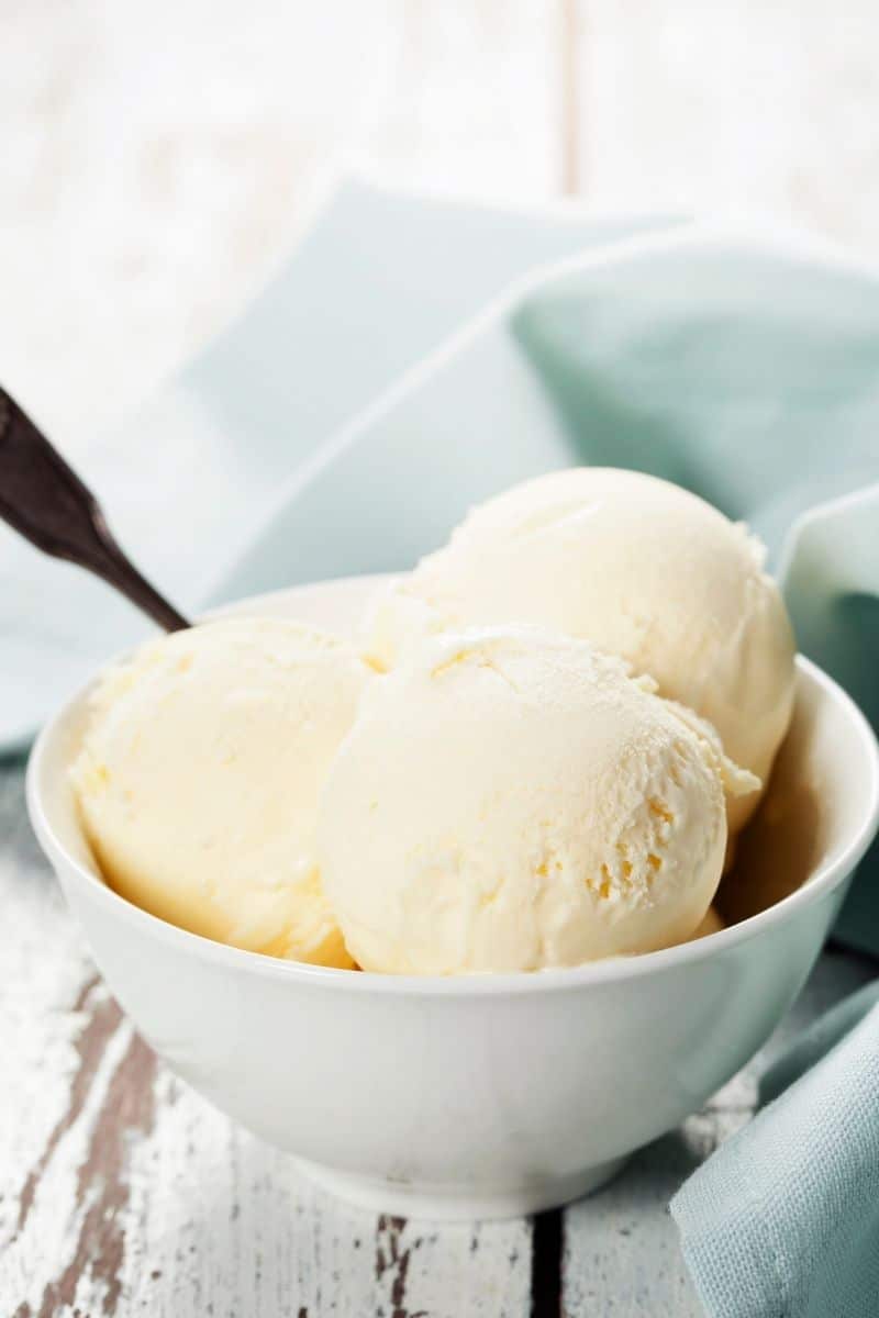 three scoops of vanilla ice cream in white bowl on distressed table with mint green napkin in background 