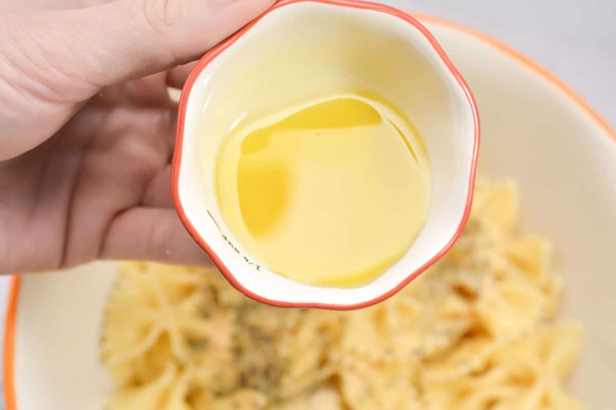 a hand holding a bowl of olive oil above a blurred bowl of pasta