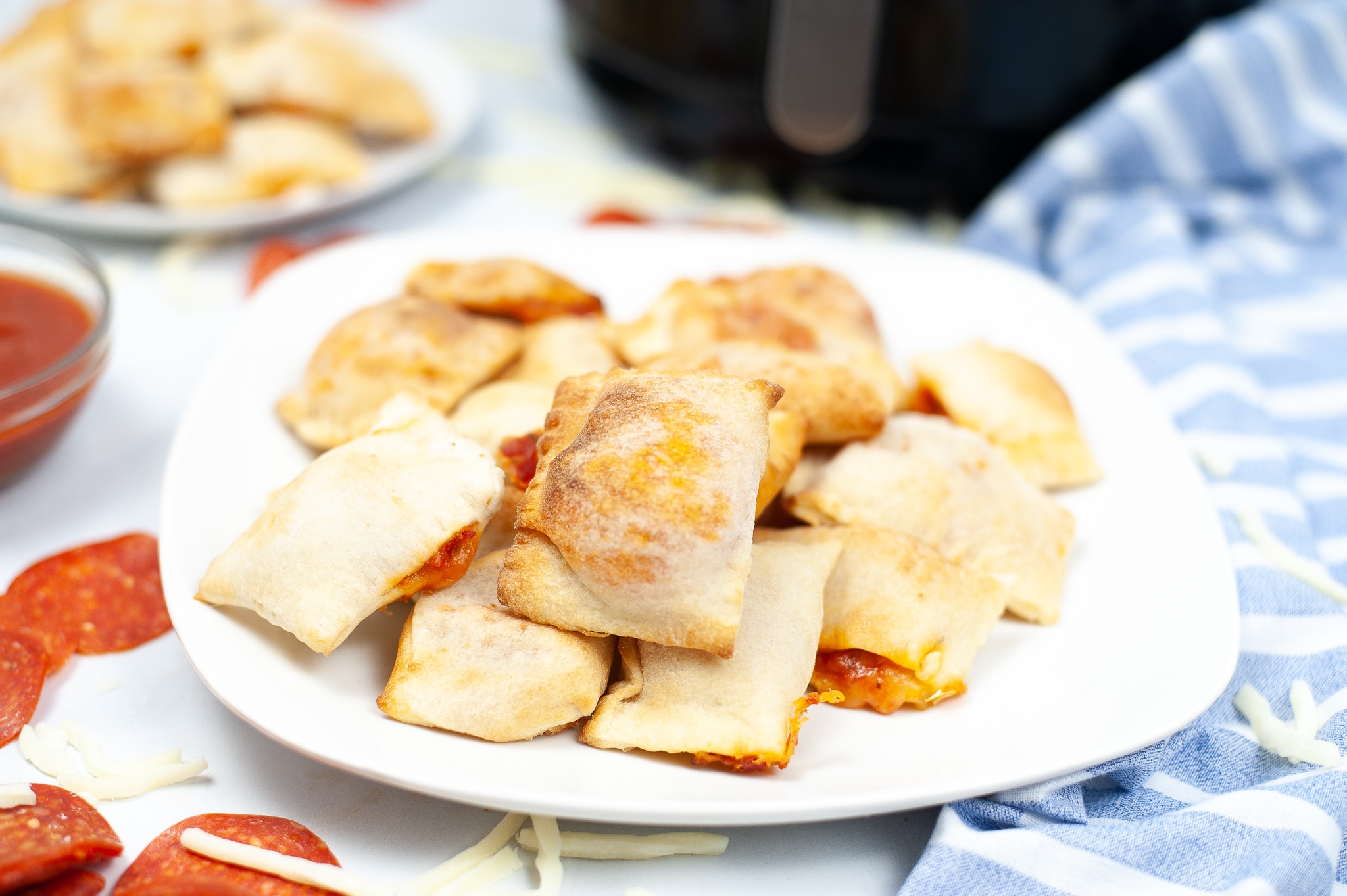 Air Fryer Pizza Rolls on a white plate with more pizza rolls and an air fryer blurred in the background 